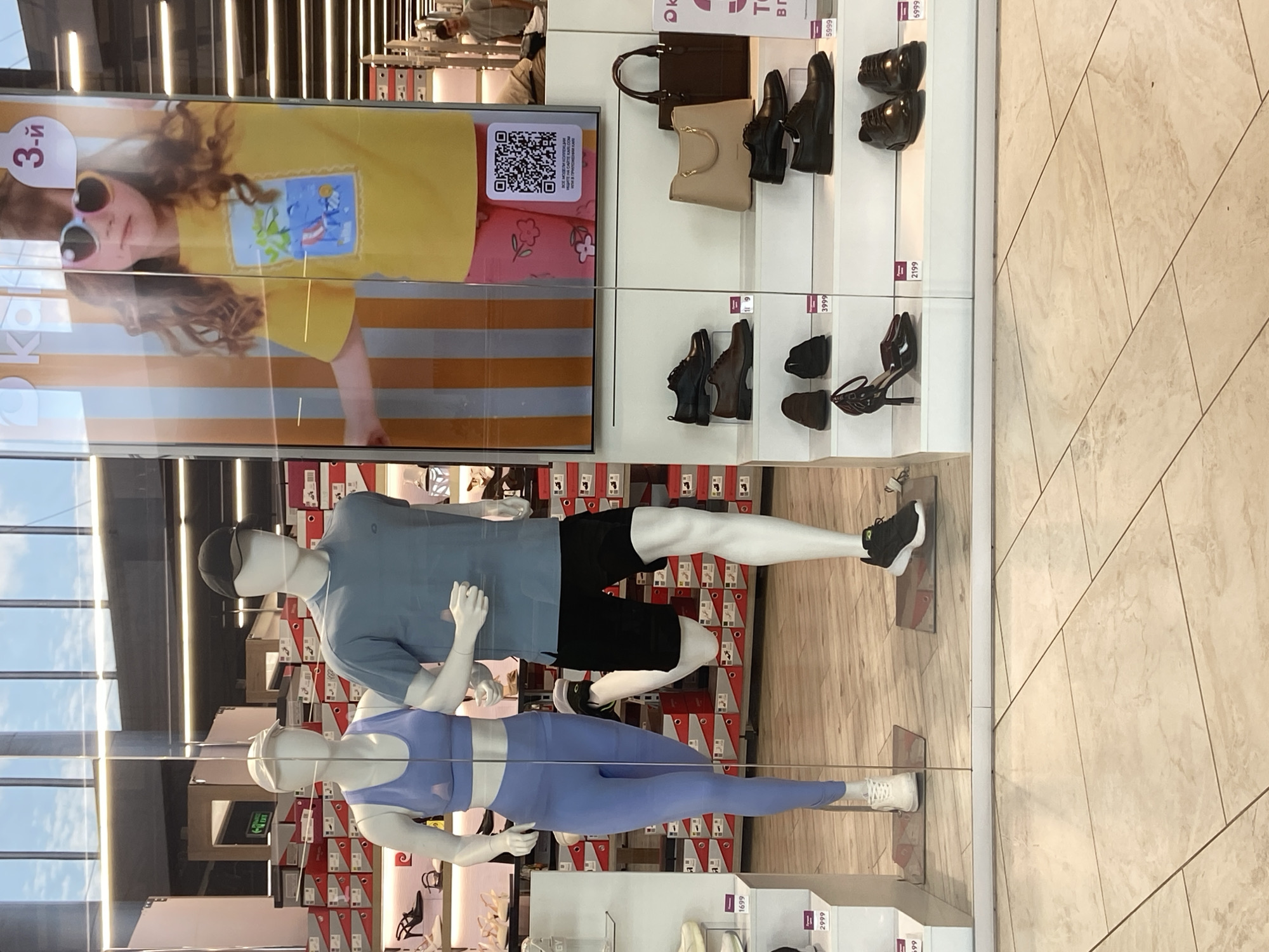 In Kirov, Russia, a shoe store window display features two mannequins, one male and one female, dressed in athletic wear and posed as if running. The male mannequin wears a blue t-shirt, black shorts, a black baseball cap, and black running shoes. The female mannequin is clad in a light blue sports bra and matching leggings, with white athletic shoes. Behind the mannequins, shelves display shoe boxes, and to the right, a large advertisement for children's clothing shows a young girl wearing a yellow t-shirt and pink shorts, with heart-shaped sunglasses. Below the advertisement, on white tiered shelving, are various men's and women's shoes, including dress shoes, loafers, and heels, as well as two handbags. The store appears to be a part of a mall or shopping center, with tiled flooring and overhead lighting visible.