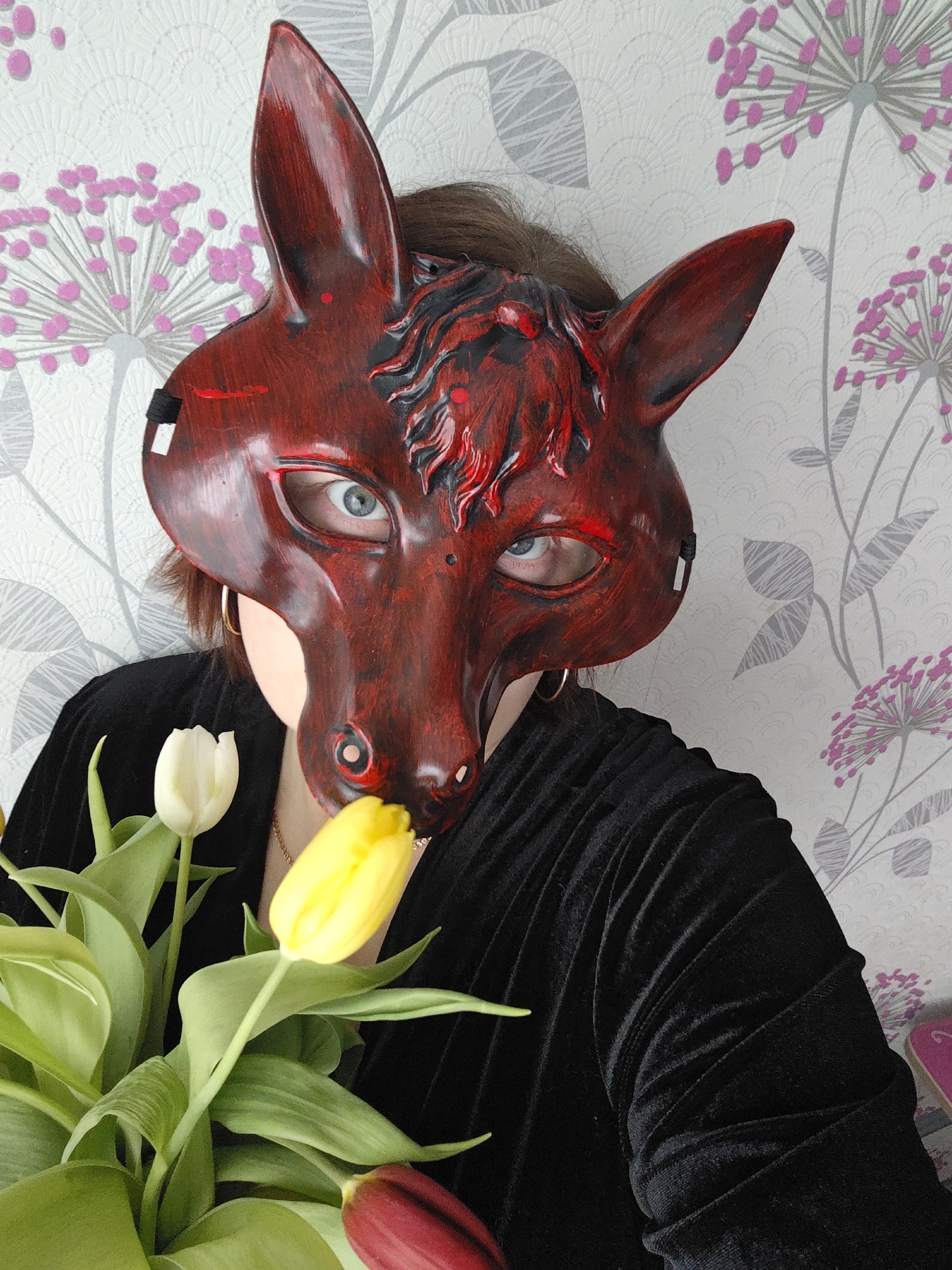 An individual is depicted indoors, possibly in Markovskii, Russia, wearing a striking red and black mask shaped like a fox or wolf head. The mask covers most of their face, revealing only their bright blue eyes, part of their chin, and some brown hair peeking out from behind the mask's ears. The mask itself is textured, with a distressed red finish and black details, including raised patterns that resemble fur or a mane. It features prominent pointed ears.
The person is holding a bouquet of tulips, prominently featuring two yellow tulips, one white tulip, and a hint of a red/darker tulip at the bottom right, surrounded by green leaves and stems. The mask's snout appears to be gently interacting with or very close to one of the yellow tulips.
The individual is wearing a black garment, likely velvet due to its soft texture and folds, and a gold chain is visible around their neck. A gold hoop earring can be seen on their right ear (viewer's left). The background consists of patterned wallpaper with abstract floral designs, resembling dandelion seed heads, in grey and pink against a lighter background. The lighting is soft and diffused, characteristic of an indoor setting. No text is visible in the image.
