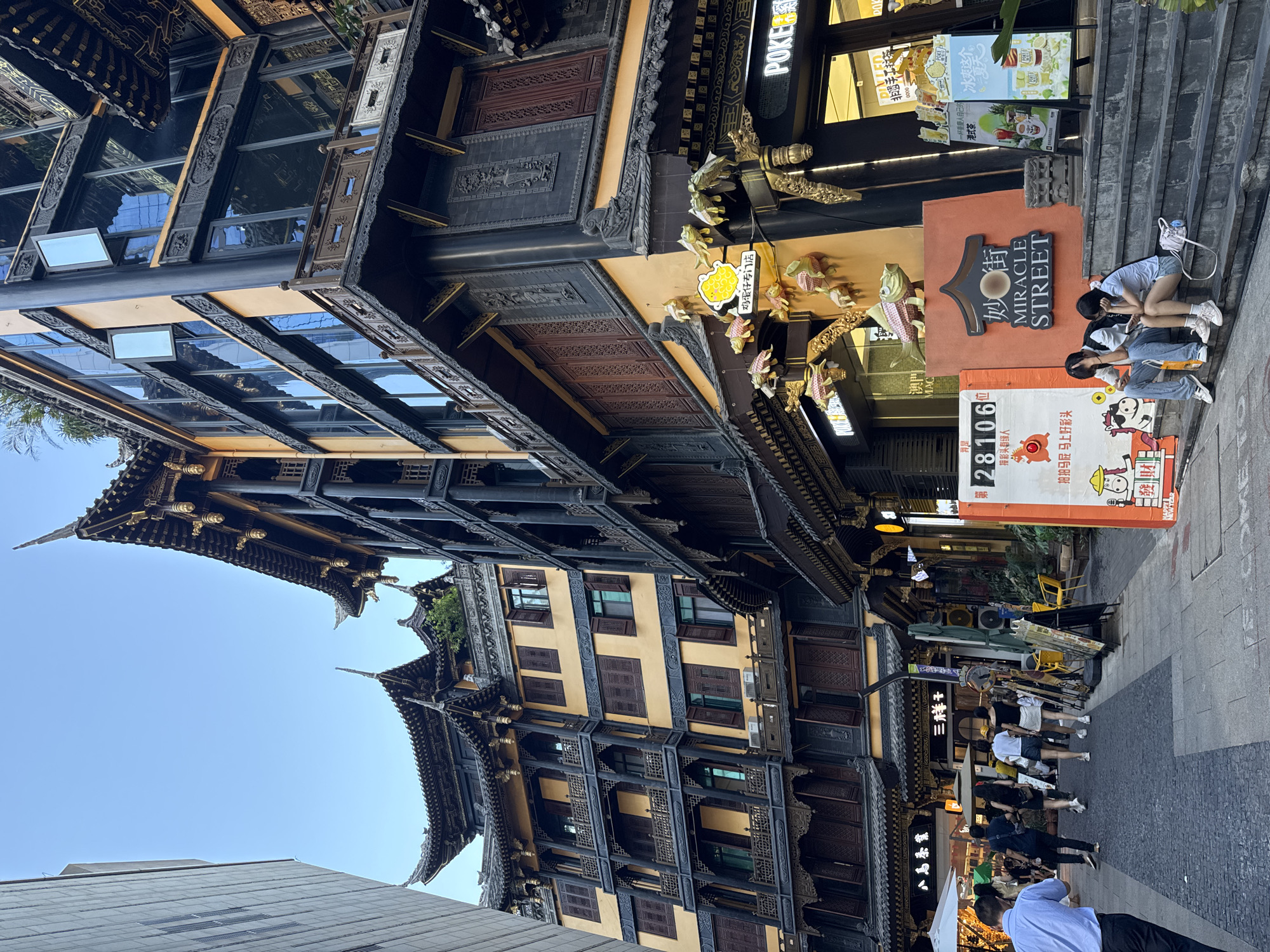 The image depicts a street scene in the Yuzhong District of China, characterized by traditional Chinese architecture with multi-story buildings featuring curved eaves and dark wooden accents. On the right, a building with a modern glass facade is integrated with the traditional design. Several people are visible walking along the paved street and seated on steps. A sign reading "MIRACLE STREET" is prominently displayed. Decorative fish ornaments hang from the building on the right. The overall atmosphere suggests a bustling commercial or tourist area with a blend of historical and contemporary elements.