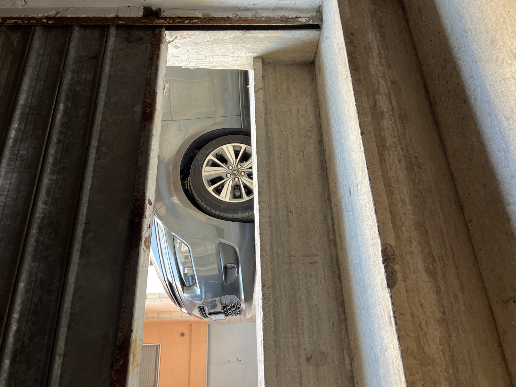 A low-angle interior view looks outward through the gap of a partially raised, dark grey, corrugated metal rolling shutter. The bottom edge of the shutter and its right vertical frame are significantly rusted. In the immediate foreground, two concrete steps, light beige and textured, rise towards the viewer, exhibiting wear and minor chipping.
Through the opening, a grey metallic vehicle, identifiable as a compact MPV or crossover, is parked on an outdoor paved surface. The front-right portion of the car is visible, featuring a distinct headlight assembly with upper LED daytime running lights, a chrome-accented grille, a parking sensor on the bumper, and a multi-spoke alloy wheel.
In the background, an orange-painted building wall with a rectangular window is visible on the left. To the right, a plain white door is present against a lighter building structure. The outdoor scene is brightly lit by natural daylight, casting shadows on the interior steps. No individuals are present. The scene is located in Santeramo in Colle, Italy.