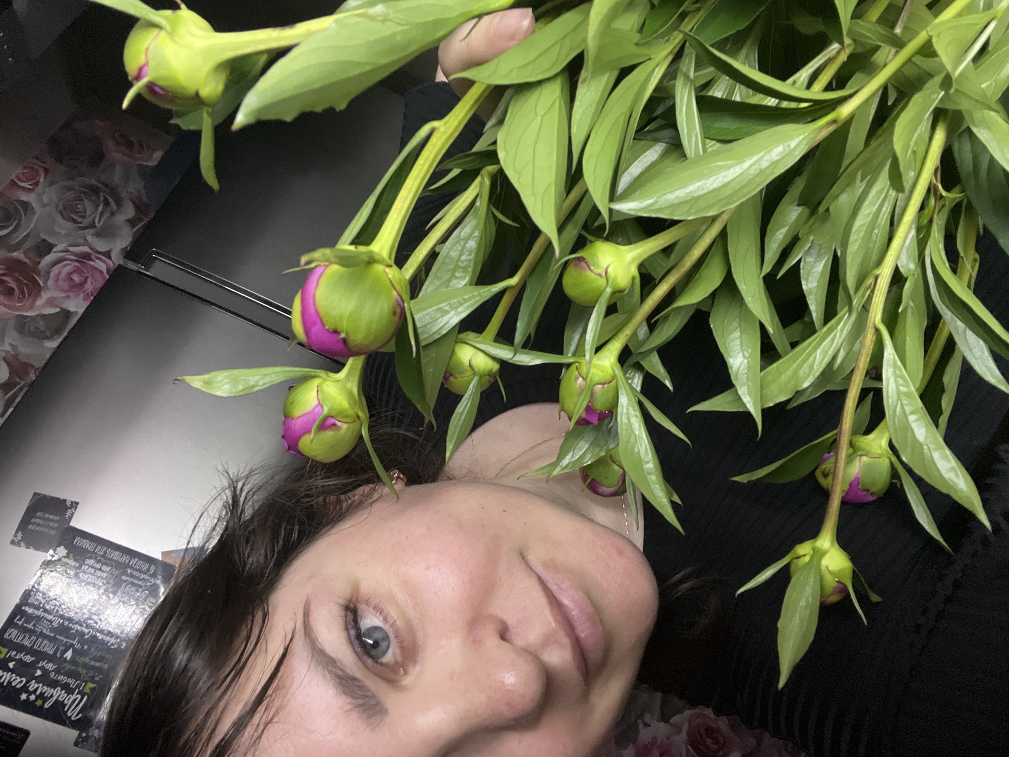 The image is a close-up, high-angle shot of a woman holding a bouquet of peonies. The woman is looking directly at the camera, and her face is partially obscured by the flowers. She has dark hair, blue eyes, and fair skin. The peonies are in various stages of bloom, with some buds and some partially open flowers. The background is dark and out of focus, but appears to be a kitchen or dining area with a refrigerator visible on the right side. There are some papers or posters on the refrigerator door. The image does not provide any information about the location in the city of Noviy, Russia.