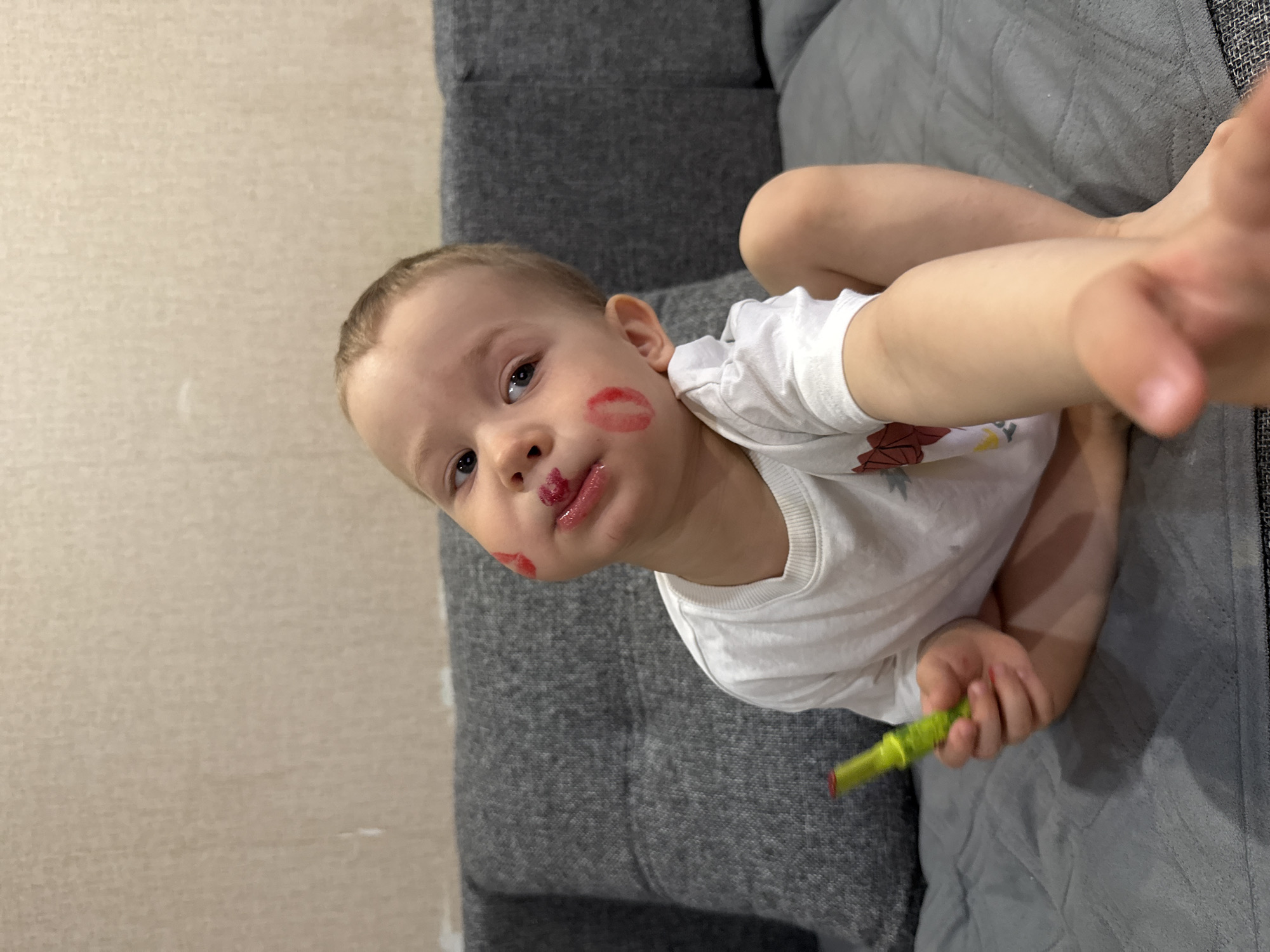 A young child is lying on a gray sofa with lipstick smeared on their face. They are holding a tube of lipstick in their hand. The location is in a residential area. I have no way to determine the city or country from this image.