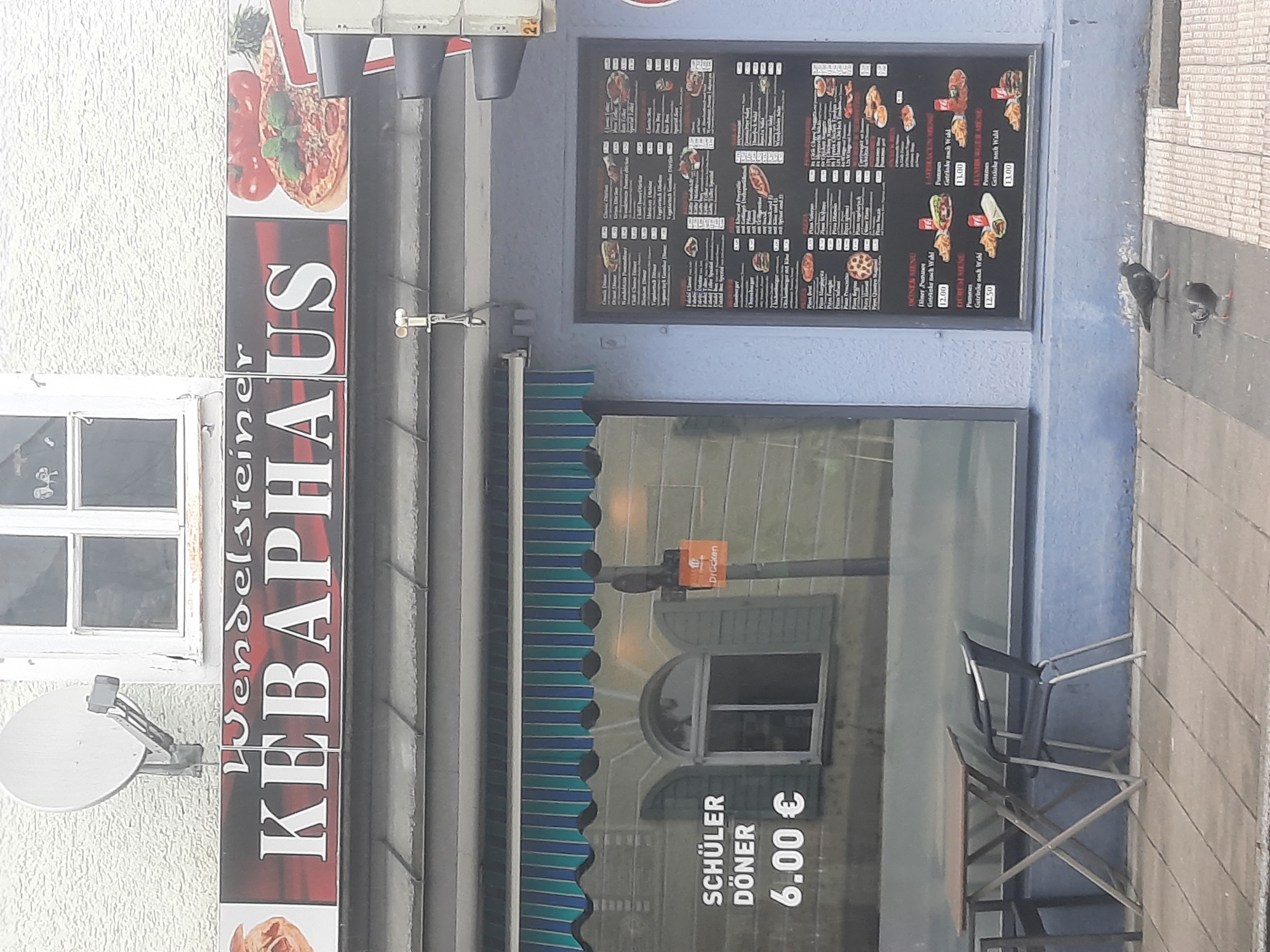 The image shows the exterior of a kebab restaurant called "WENDELSTEINER KEBABHAUS" located in Rosenheim, Germany. The restaurant has a blue facade with a large window. On the left side of the window, there is a sign that reads "SCHÜLER DÖNER" with a price of "6.00 €". Inside the window, a table and chairs are visible. To the right of the window, a menu is displayed on the facade. The menu lists various food items including döner, pizza, and hamburgers. The street in front of the restaurant is paved with tiles. Two pigeons are standing on the pavement. The building has a window and a satellite dish on its upper level. A traffic light is partially visible on the right side of the image.