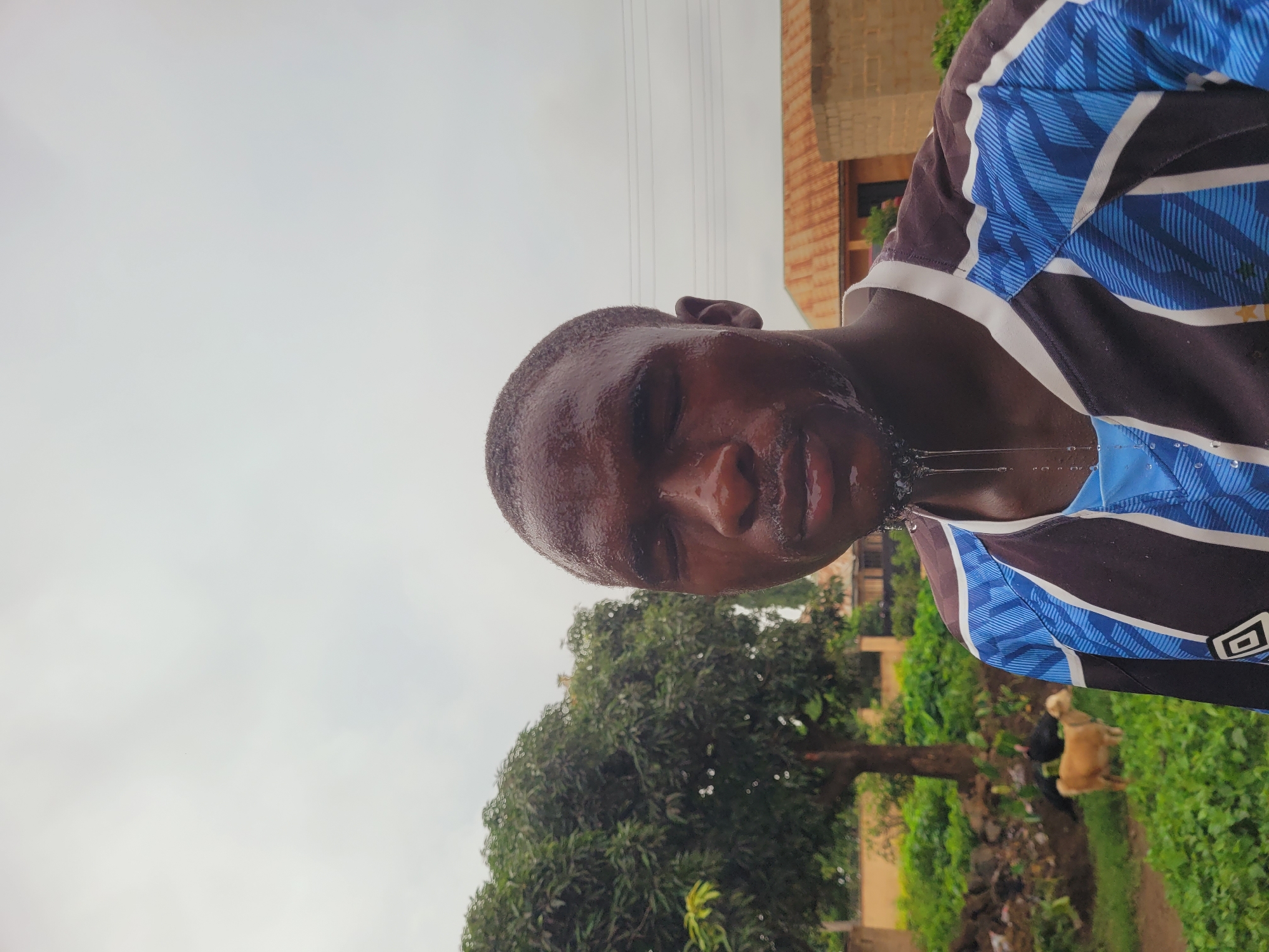The image, taken in Bukuru, Nigeria, is a portrait of a Black man from the chest up. He is wearing a blue and black striped shirt. His face is wet with sweat or water, and droplets are visible on his forehead and chin, with some falling onto his shirt. His eyes are closed, and his mouth is slightly open, suggesting he may be drinking or spitting. The background is blurred and consists of greenery, a tree, a building, and a hazy sky. The image is oriented vertically, with the man's head appearing at the bottom and his shoulders at the top, indicating it was taken in landscape orientation and then rotated.