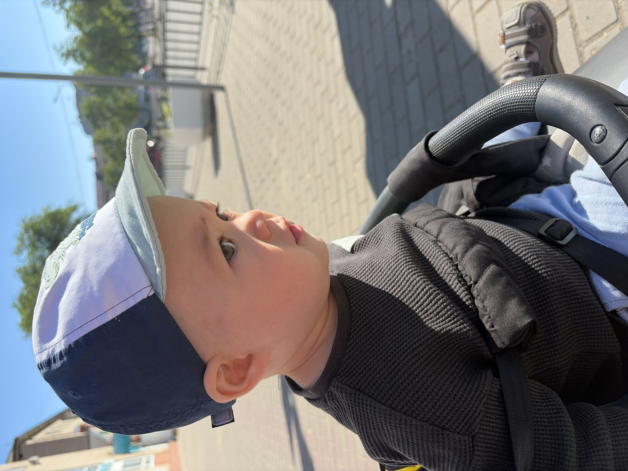 A young child wearing a navy blue and light blue baseball cap and a dark gray textured long-sleeved shirt is seated in a stroller. The child is looking to the right with their mouth slightly open. The stroller has black handlebars and a black harness with a blue strap visible. A small portion of the child's light blue pants and a brown sandal are also visible. The background shows a paved area, some buildings, trees, and a bright blue sky. The scene appears to be in a city environment, possibly Chaikovsky, Russia, given the context.