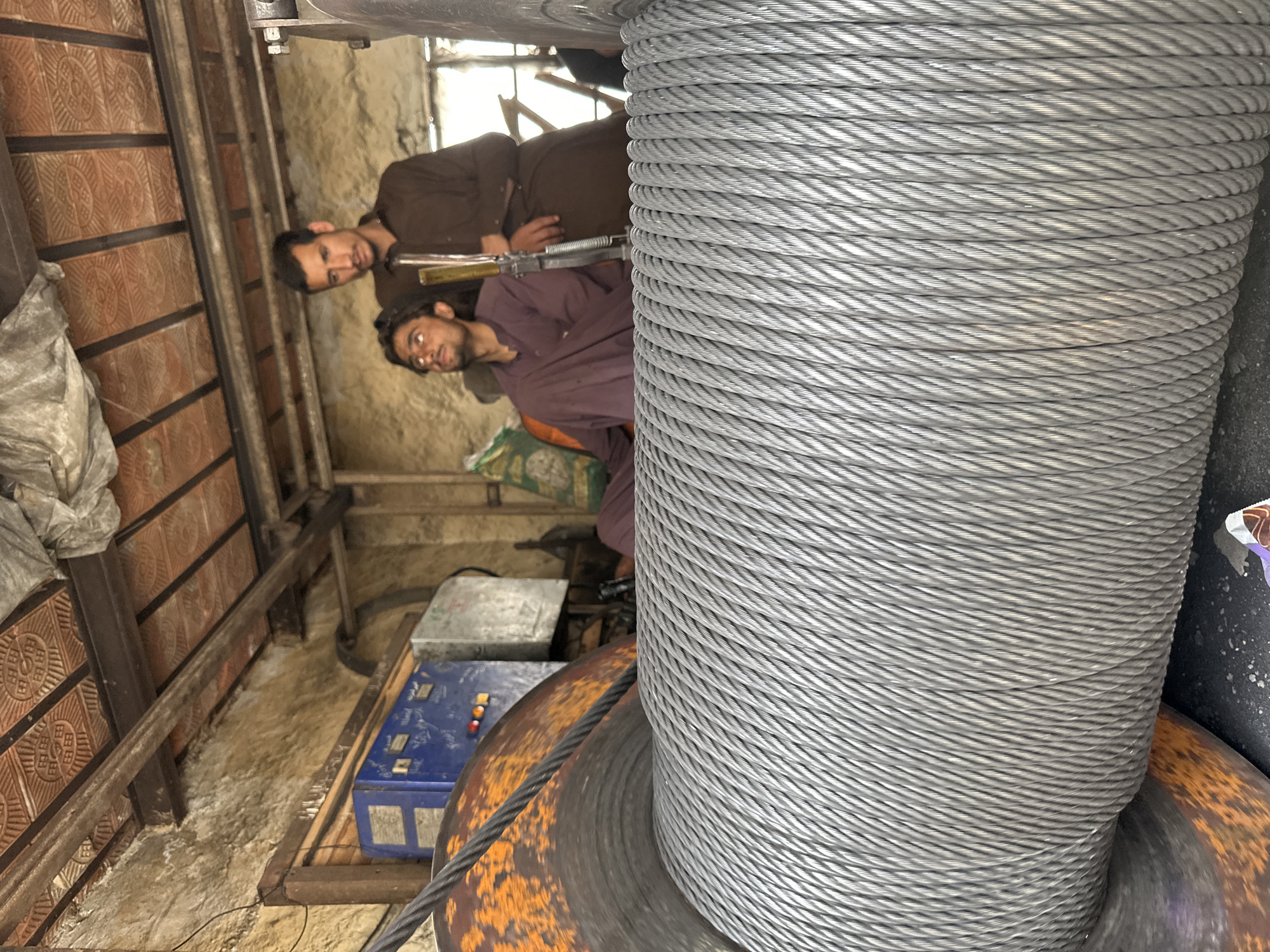 In Quetta, Pakistan, a close-up shot captures a large spool of galvanized steel cable in the foreground, filling most of the frame. The cable is tightly wound, with individual strands visible and catching the light. Behind the spool, two young men are partially visible in a workshop or industrial setting. The man on the left, wearing a purple shalwar kameez, looks upwards with his head tilted back. The man on the right, in a brown shalwar kameez, stands behind him and looks towards the camera. The background reveals a rustic environment with a corrugated metal ceiling, exposed wooden beams, a blue electrical control panel with red and green lights, and a rusted orange metal component of the spool's mechanism. The overall impression is one of a working industrial space.