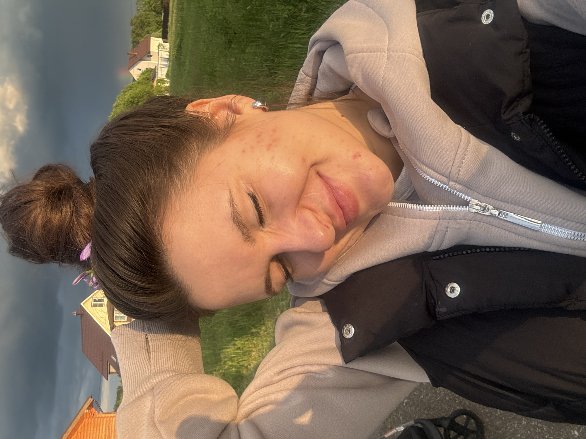 A sideways view of a woman lying on grass, with houses visible in the background. The woman has brown hair in a bun, adorned with purple flowers, and she wears a light-colored hoodie and a black vest. Her eyes are closed, and she appears to be smiling slightly. The location is inferred to be Vysokinichi, Russia based on other information not present in the visual image. The background shows a green field, houses with orange roofs, and a sky with ominous dark clouds. A rainbow is barely perceptible in the background. A stroller is present in the very lower corner of the image.