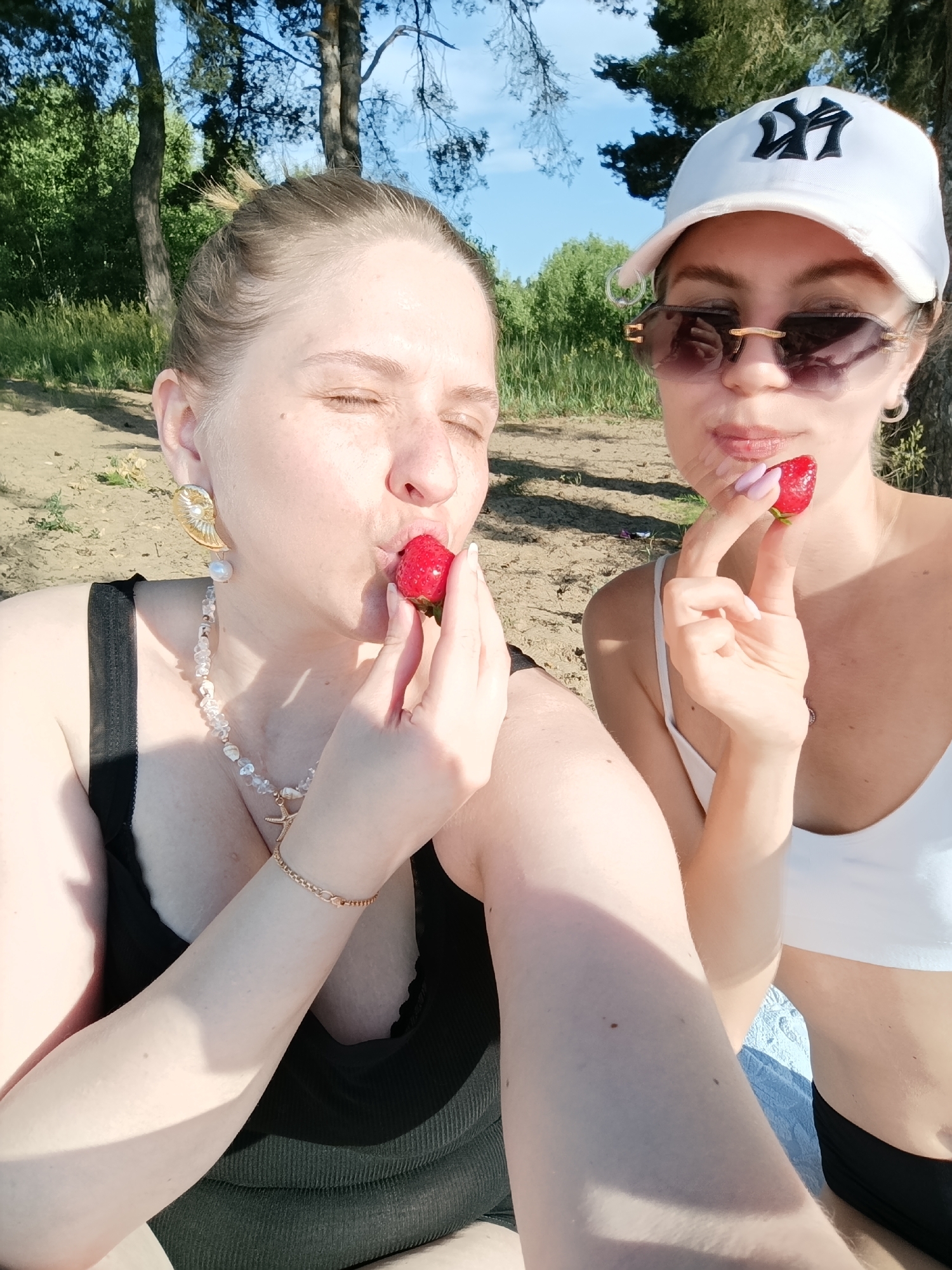 Two young women are pictured outdoors, seated on a sandy area near trees. The woman on the left, with her eyes closed and wearing a black top and a pearl and seashell necklace, is bringing a strawberry to her mouth. The woman on the right, wearing a white crop top and a white baseball cap, is holding a strawberry and appears to be about to eat it. She is also wearing sunglasses and hoop earrings. The background consists of green foliage and trees, with a bright blue sky overhead. The lighting suggests it is a sunny day. The location is identified as Podstёpki, Russia, likely a park or recreational area given the natural setting.