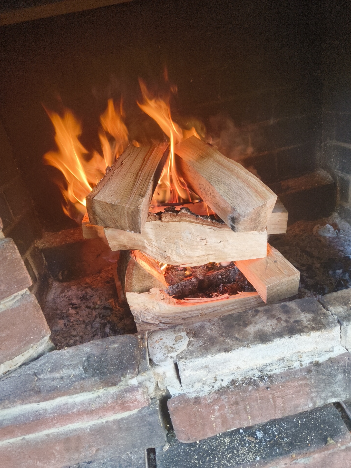 The image depicts a fireplace with a fire burning. Several logs of wood are stacked within the fireplace, with flames engulfing the lower logs and rising upwards. Embers and ash are visible beneath the burning wood. The interior of the fireplace appears to be constructed of bricks, with some mortar visible between them. The background is dark, suggesting the interior of a room or a darkened area behind the fireplace. There are no people visible in the image. This scene is consistent with a typical fireplace in a residential setting, potentially in Востец Снт, Russia, although the specific location cannot be definitively confirmed from the image alone.