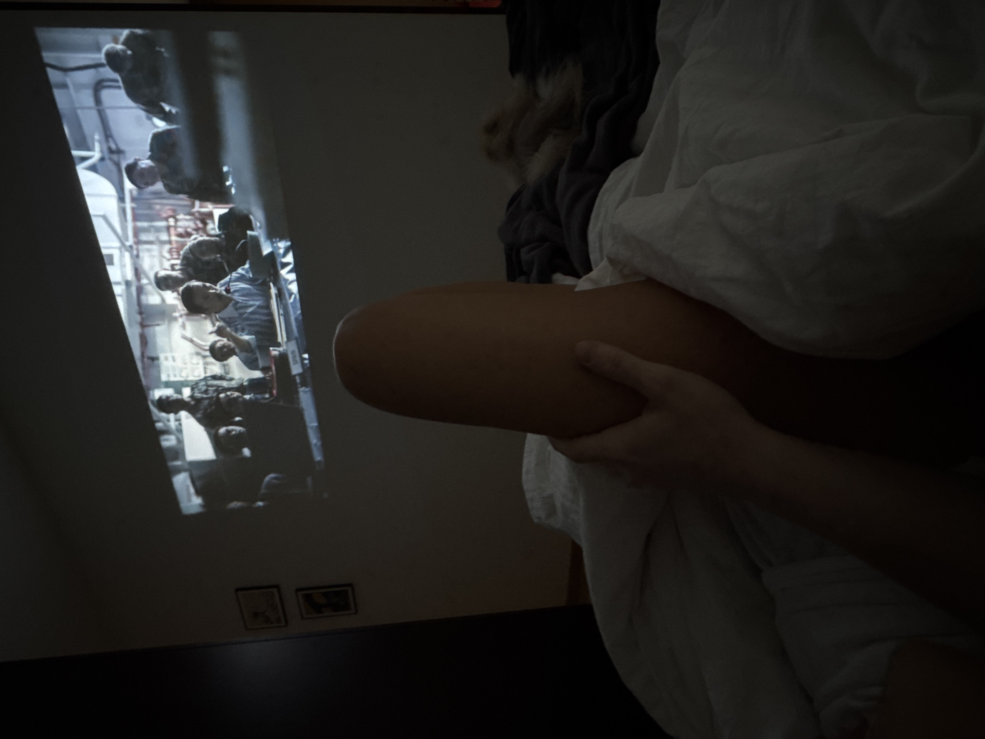 A dimly lit indoor scene shows a person seated and covered with a white blanket. Part of their leg is visible, and their hands are clutching it. On the wall to the left, a projected image depicts a group of people in what appears to be a submarine interior. Below the projected image are two small, square pictures. The floor is dark, adding to the overall low light conditions.