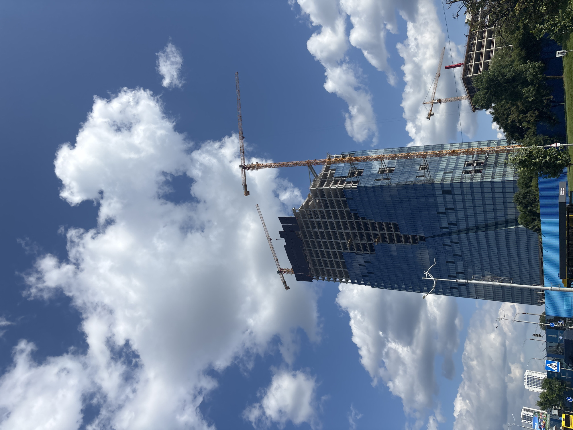 The image captures a construction site in Minsk, Belarus, dominated by a partially completed skyscraper with a reflective blue glass facade. Two large yellow tower cranes are actively engaged in construction, one prominently positioned next to the skyscraper and another to its right, serving a lower building also under construction. The sky is a vibrant blue, dotted with fluffy white cumulus clouds. In the foreground, a street with traffic lights and pedestrian crossings is visible, along with blue construction fencing and greenery. The scene depicts urban development under a bright, sunny sky.