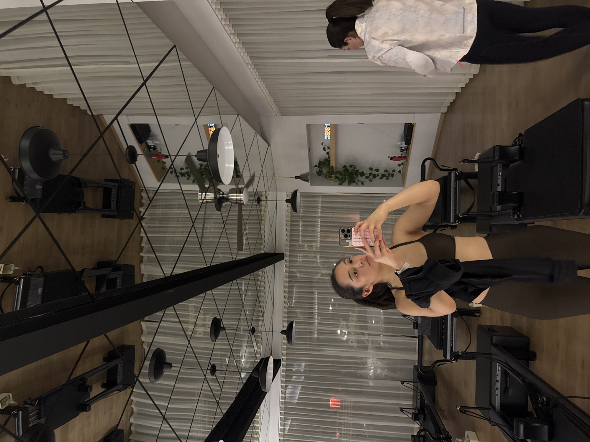A dimly lit indoor space with mirrored walls and a high ceiling filled with linear lighting fixtures and decorative pendant lamps. A woman in fitness attire holds a mobile phone to take a picture, while another woman stands in the upper-right corner of the frame wearing a light jacket and dark pants. Exercise machines, possibly Pilates reformers, are arranged in rows on the wooden floor. The location appears to be a fitness studio in Puebla, Mexico.