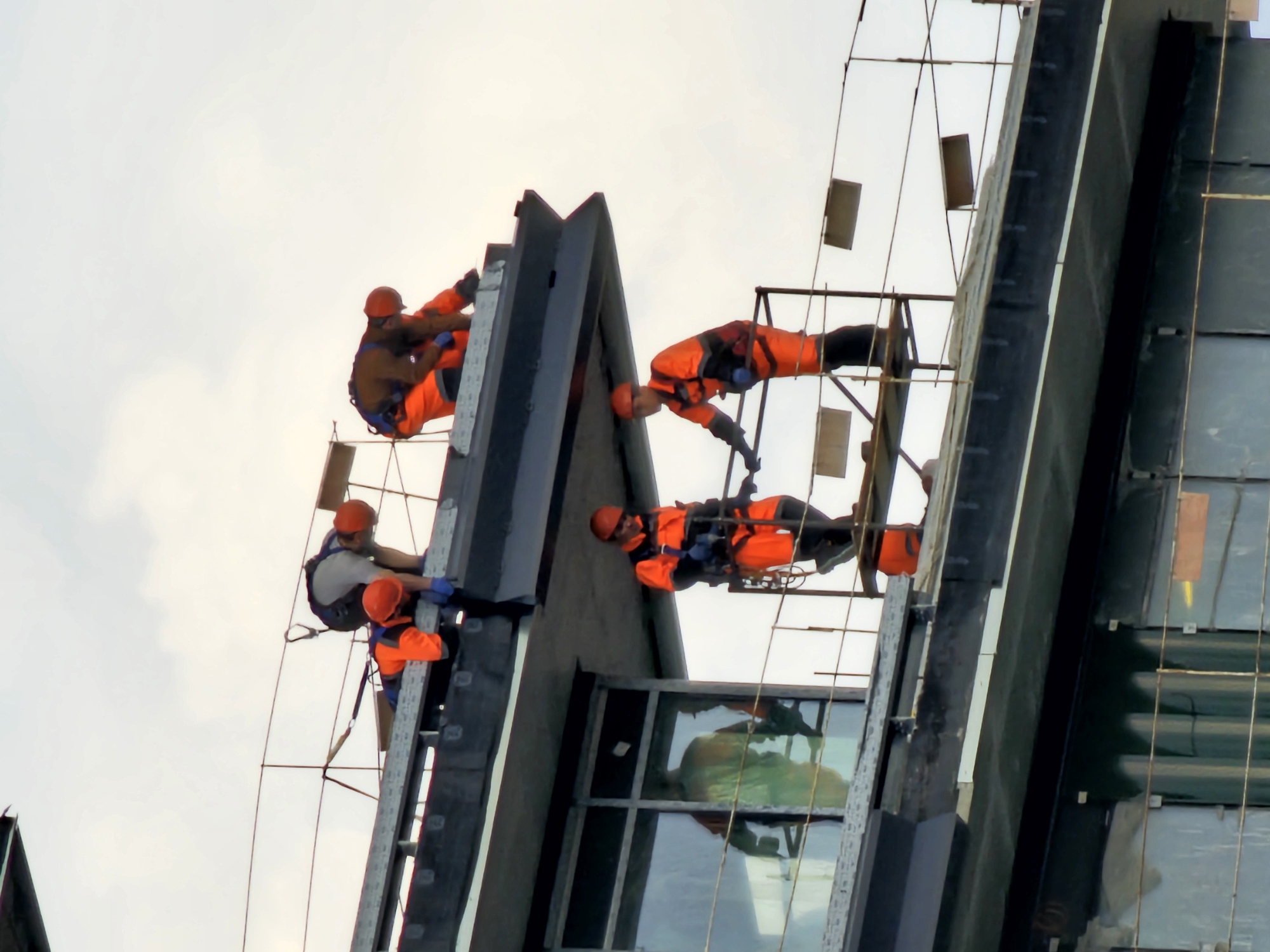 Several workers are performing maintenance on a skyscraper in Moscow, Russia. They are wearing orange safety vests and hard hats, and they are suspended from ropes and scaffolding along the exterior of the building. The building has a modern design with glass windows and dark gray panels. The sky is overcast.