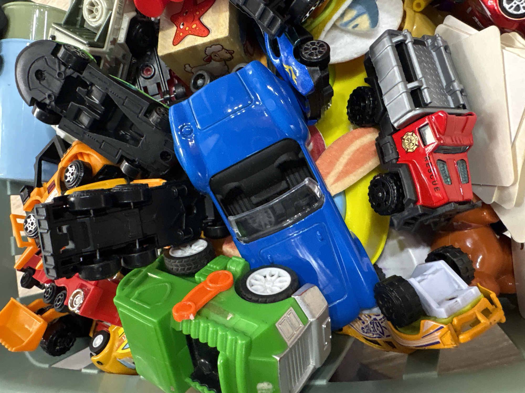 A close-up, overhead view shows a pile of colorful toy cars of various sizes and types. Dominating the center is a bright blue toy sports car, flipped on its side. To its left, a large green toy garbage truck is also on its side, with a visible orange plastic lever. A black toy car with a detailed undercarriage is positioned on top of the green truck. Several other toy vehicles, including a red fire truck and a yellow car, are scattered throughout the frame, some partially obscured. The toys are packed into a container, the edges of which are visible. The background is blurred, with hints of other toys and containers. The image is well-lit, with no discernible shadows. There are no people or animals visible in the image. The scene is static, with no actions or interactions taking place. The location in Mikhaylovsk, Russia, is not discernible from the image itself, as it depicts a collection of toys in a container and provides no contextual clues about its geographic location.