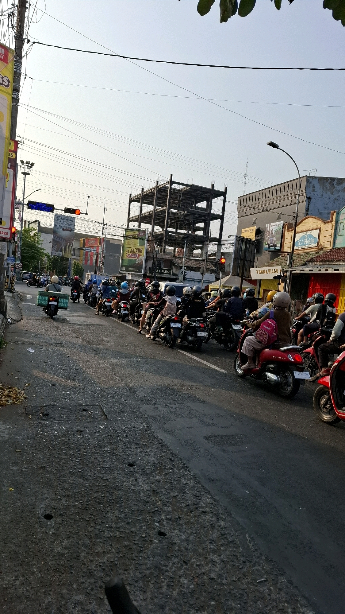 The image depicts a street scene in Damaran, Indonesia, characterized by a significant traffic jam of motorcycles. The perspective is from a low angle, looking forward along a street lined with buildings and construction. The sky is clear with some overhead wires. In the foreground, the asphalt road shows signs of wear and repair. A long line of motorcycles and their riders are stationary or moving slowly in traffic. Many riders are wearing helmets, and some are carrying passengers or goods. Buildings are visible on both sides of the street, some appearing to be shops or commercial establishments, with one building under construction featuring a prominent metal frame. Traffic lights are visible, indicating a signalized intersection. The overall impression is one of congested urban traffic in an Indonesian city.