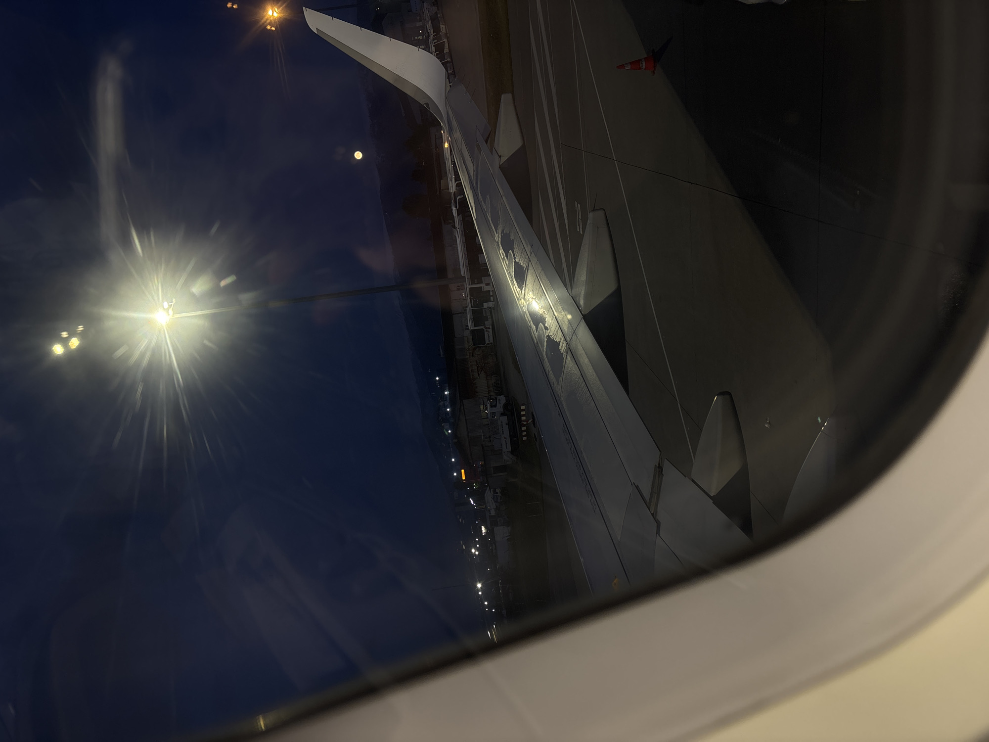 The image depicts an airplane wing viewed through an airplane window at night. The aircraft appears to be on the ground, likely at an airport, given the presence of runway markings, lights, and what appears to be an orange traffic cone. The reflection in the window shows streetlights and possibly buildings in the background. The sky is dark, with what could be the moon visible.