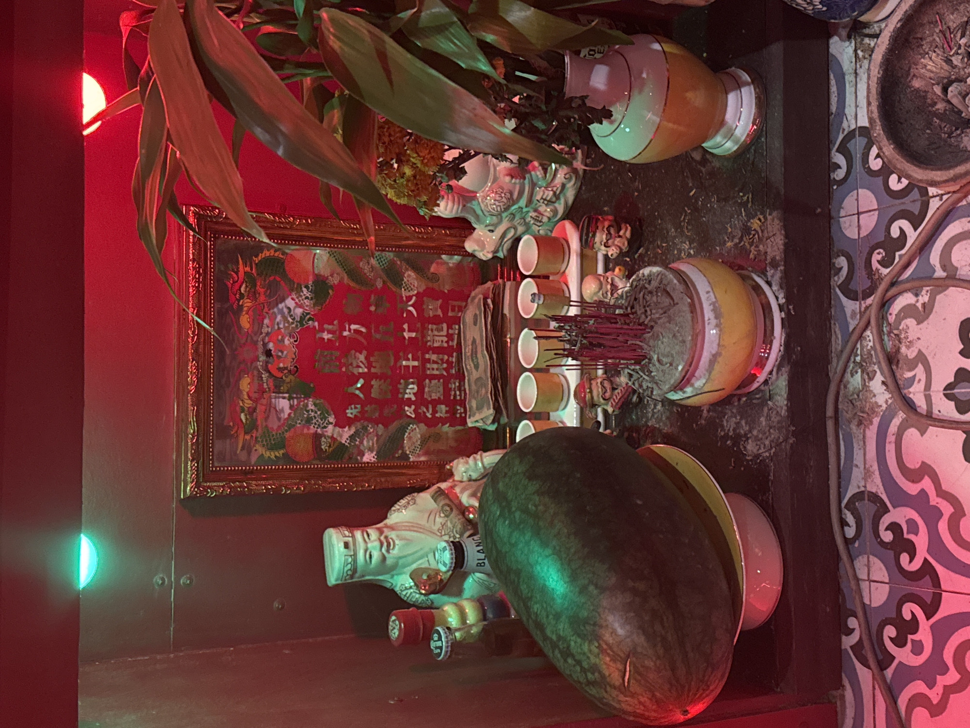 This photograph shows a small Buddhist altar, likely in a home or small business in Đà Nẵng, Vietnam. The altar is crowded with various religious objects and offerings. A large, ripe watermelon sits prominently on a plate in the foreground. Behind it is a statue of an elderly Chinese god figure holding a bottle, possibly representing longevity or prosperity. Several smaller bottles and cans are arranged around the statue. Above these objects are a row of small cups or vessels and a pot filled with incense sticks, some of which are lit and emitting smoke. A gold-framed picture with red coloring and Chinese characters hangs behind the altar, with further ornaments and plants on top. The altar is placed against a red wall, while the floor or surrounding surface features a mosaic tile pattern in white, blue, and purple. The scene has a mix of cultural and spiritual symbolism, reflecting Vietnamese Buddhist practices of honoring ancestors and seeking good fortune.