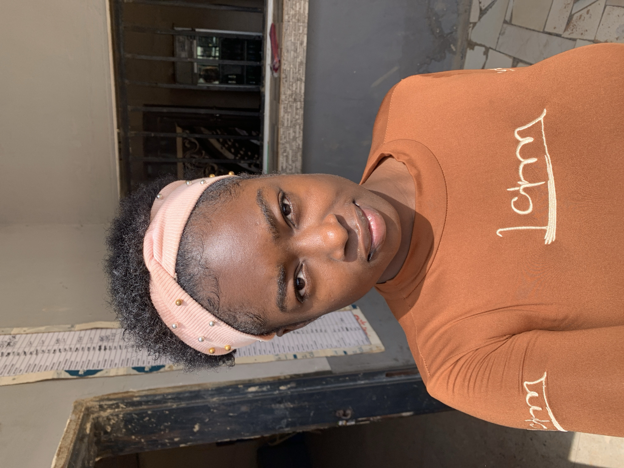 A young woman with dark skin, short black hair, and a light pink headband with small gold and silver studs is posing for a portrait. She wears a brown long-sleeved shirt with white cursive lettering. She appears to be standing outside of a building. A window with iron bars is behind her. A white wall, a sign with text, and a dark-colored wooden door are also visible. The image was taken during daylight hours. No information confirms the location is in New Stadium, Nigeria.