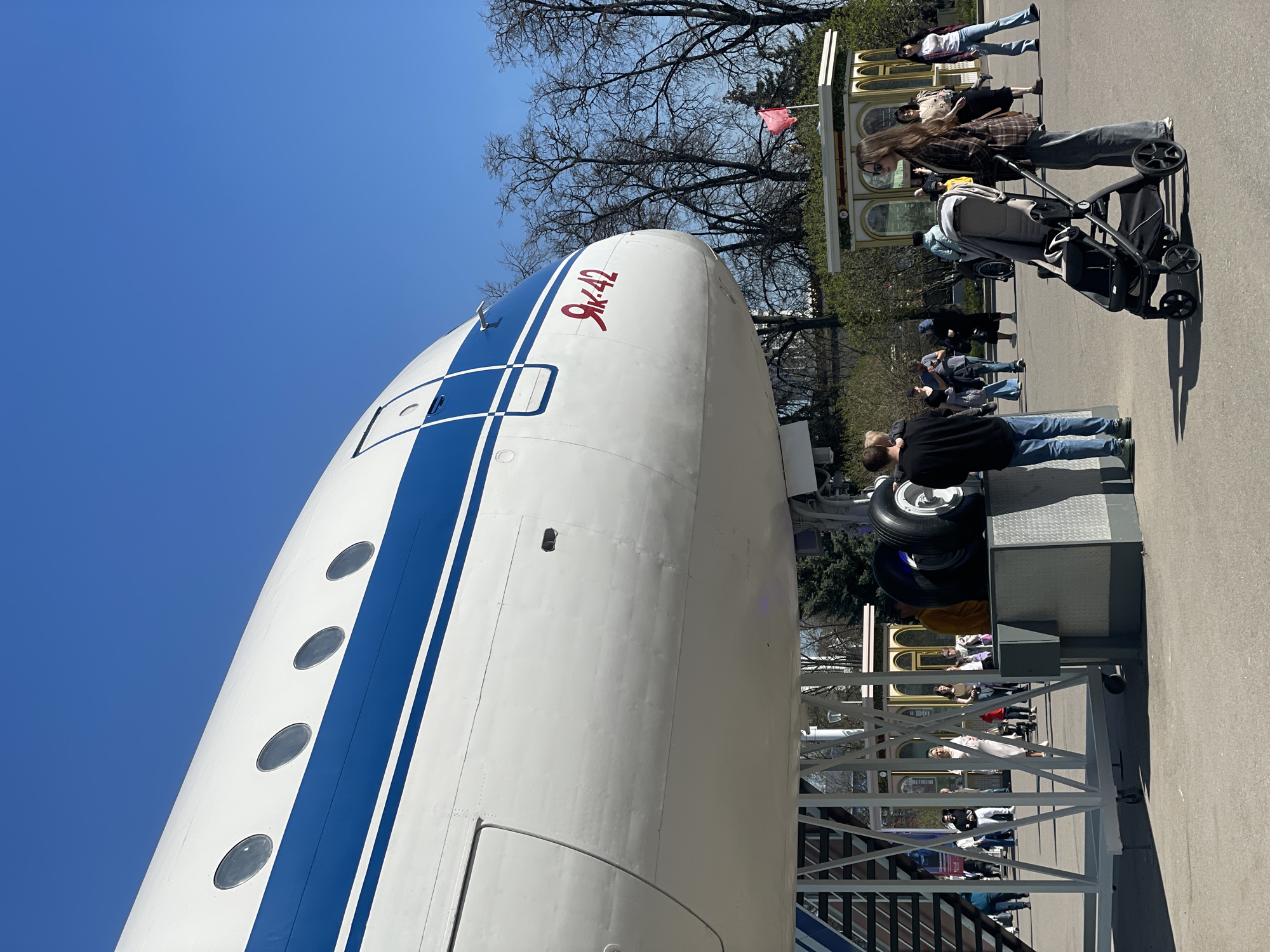The image captures the nose of an Ilyushin Il-62 aircraft, designated "9k-42" in Cyrillic lettering. The aircraft is white with a broad, blue stripe running along its length, punctuated by a row of circular windows. A blue sky fills the background above the aircraft.
The aircraft is stationed on a platform with attached stairs for public viewing. Several people are visible in the surrounding area. To the right, a woman pushes a stroller, while other individuals stand and stroll along a paved area. In the background, there is a decorative structure, possibly a kiosk or building facade. Bare trees are also visible, suggesting it may be late autumn or early spring. The location is potentially a park or museum where the aircraft is on display. The city and location are not identifiable from the image.