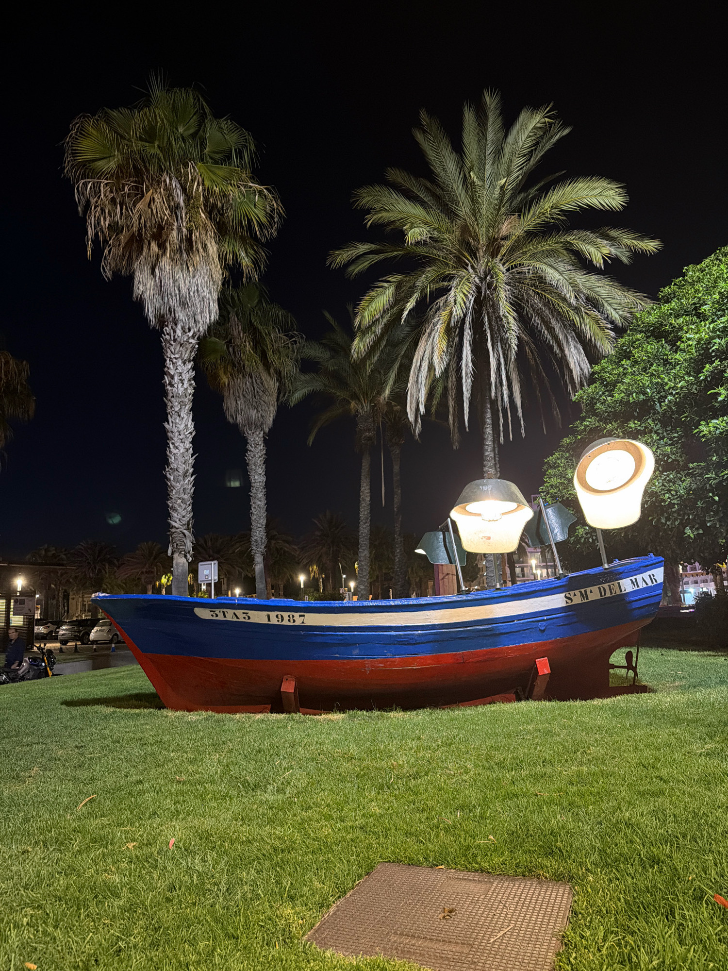 The image depicts a brightly painted blue and red boat placed on a grassy area at night in the city of Salou, Spain. The boat is oriented with its bow to the left and stern to the right. It has white lettering on its side that reads "STA3 1987" and "SM DEL MAR". Two large, inverted lamps are mounted on the boat, emitting a warm light.
Several palm trees are visible in the background, their fronds illuminated by artificial light. The sky is dark, indicating it is nighttime. To the left of the boat, a person is seated on a scooter or motorcycle. In the mid-ground, several cars are parked. The ground in the foreground is covered with well-maintained green grass and a metal grate.
The scene appears to be an outdoor public space, possibly a park or promenade. The overall impression is of a decorative installation or a landmark within the city.