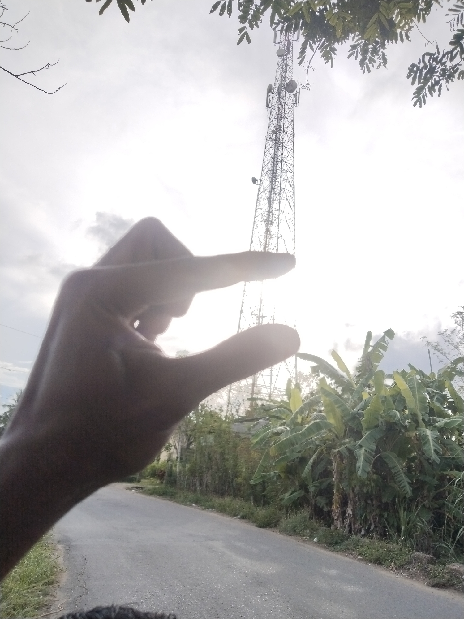 A photo taken on a road in Lhokseumawe, Indonesia, shows a hand pinching a telecommunications tower between its thumb and index finger. Banana trees and dense vegetation are visible on the side of the road. The sky is overcast.