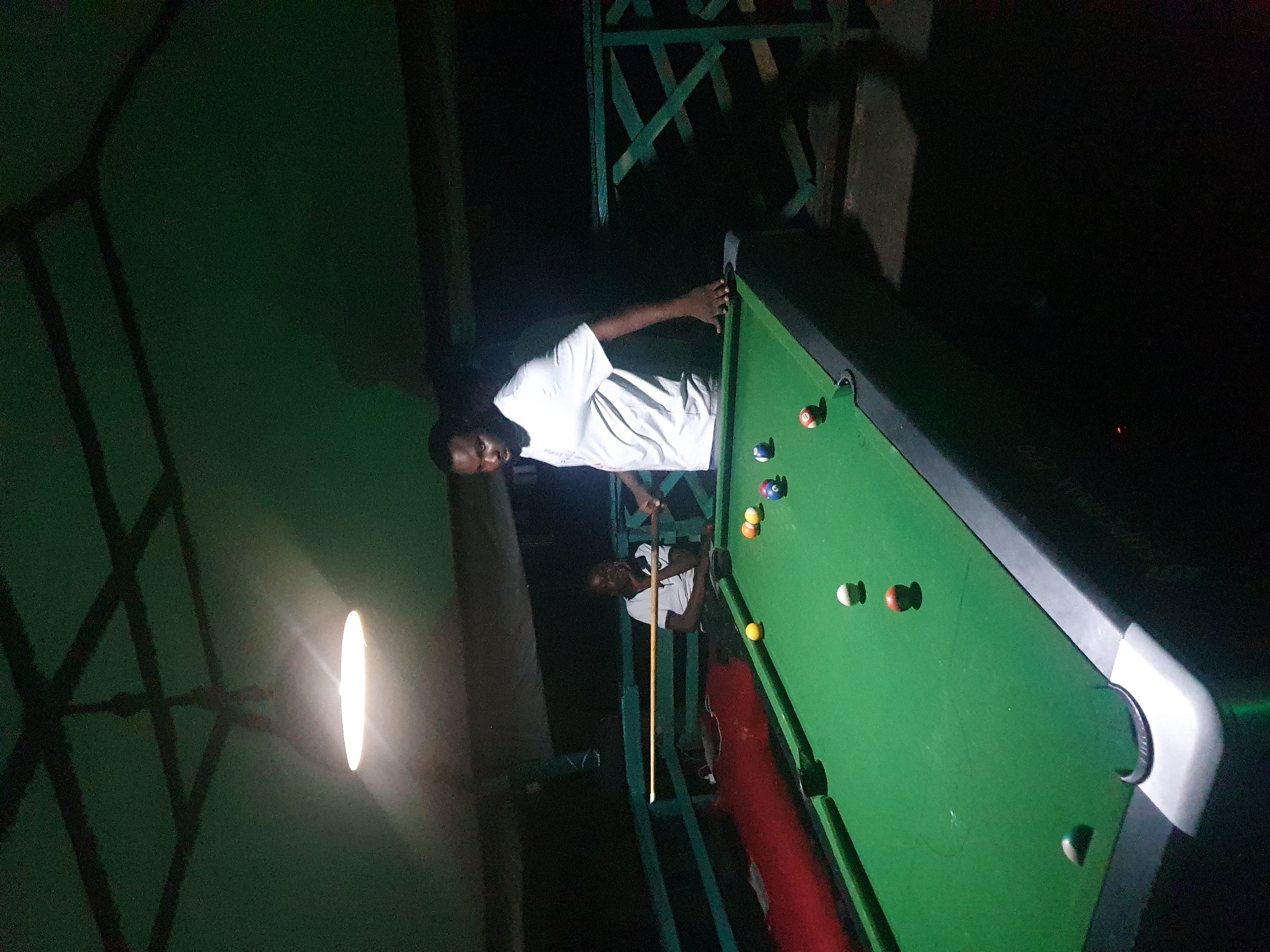 A dimly lit outdoor billiards scene in Lokoja, Nigeria, features two individuals engaged with a pool table. A man in a white t-shirt stands to the right of the table, his hand resting on the edge, poised as if to play. To the left, a second individual, also in a white t-shirt, is seated, looking towards the table with their chin resting on their hand. The pool table is green with several colored balls scattered across its surface. Overhead, a light fixture illuminates the area, casting shadows. The background reveals a green railing and dark, unidentifiable structures. The overall impression is of a casual, nighttime gathering for a game of pool.