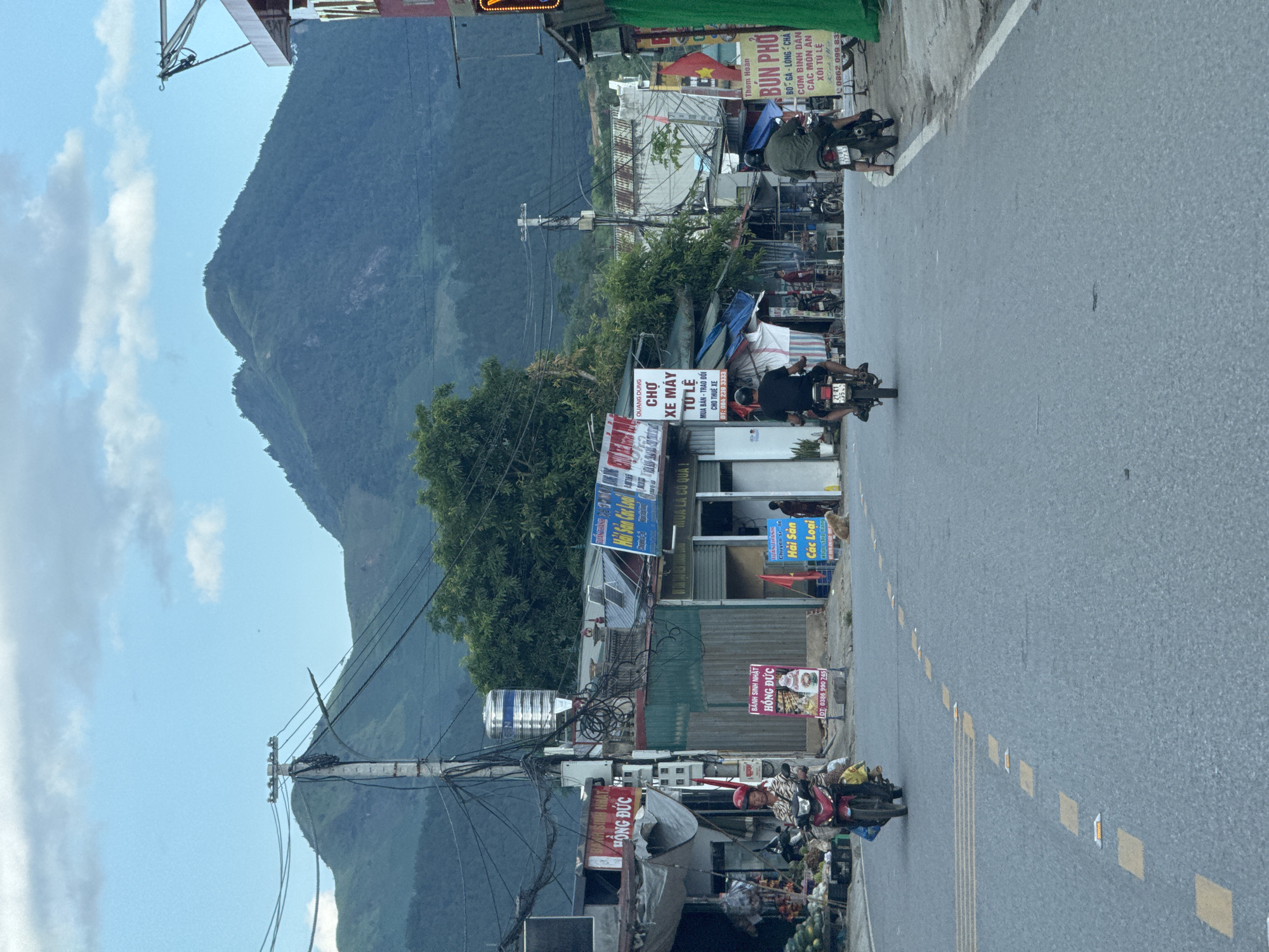 The image captures a street scene in Tú Lệ, Vietnam, characterized by a paved road leading towards a backdrop of prominent green mountains under a bright blue sky with scattered white clouds.
In the foreground and midground, the asphalt road displays yellow dashed lines and reflective road markers. Three individuals on motorbikes are visible: one on the left in a red helmet and a striped shirt, riding a red and black scooter, and two further down the road, seen from behind, on darker motorbikes.
The sides of the road are lined with a mix of single-story buildings and makeshift structures, predominantly commercial. Signs in Vietnamese are prominent. On the left, a stall displays various fruits and produce, including watermelons, next to a building with a "BÔNG ĐỨC" sign and a "HẢI SẢN CÁC LOẠI" (Seafood of all kinds) sign. A red sign for "BÁNH SINH NHẬT HỒNG ĐỨC" (Hồng Đức Birthday Cake) with a phone number is also visible.
In the mid-ground, a sign clearly states "CHỢ XE MÁY TÚ LỆ" (Motorbike Market Tú Lệ) with "MUA BÁN - TRAO ĐỔI CHO THUÊ XE" (Buy - Sell - Trade - Rent Motorbikes) and a phone number (084 239 3333). Another large sign on the right advertises "BÚN PHỞ BÒ - GÀ - LÒNG - CHẢ CƠM BÌNH DÂN CÁC MÓN ĂN XÔI TÚ LỆ" (Bun Pho Beef - Chicken - Sausage Rice Dishes various dishes sticky rice Tú Lệ) with a phone number (0862 099 833). Vietnamese flags are affixed to some structures.
Utility poles with dense, tangled networks of overhead electrical and communication wires are a dominant feature. One pole structure has a large, cylindrical stainless steel water tank mounted on it. Trees and dense foliage are visible behind and between the buildings, blending into the base of the mountains. A light brown dog is observed sitting on the sidewalk on the right side of the road.