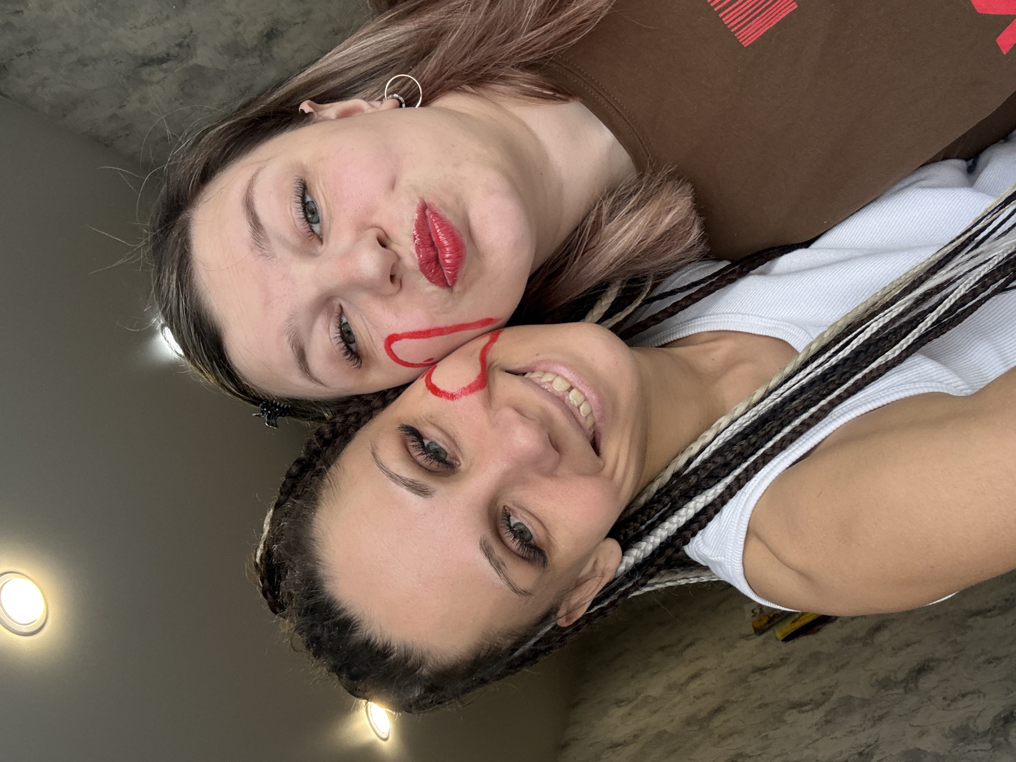 Two young women are captured in a close-up, rotated portrait, seemingly taken from a low angle looking upwards, possibly while they are lying down. The setting appears to be indoors with a muted, concrete-like wall or ceiling in the background, illuminated by two circular recessed lights.
The woman at the top (left in the rotated image) has fair skin, light brown hair with blonde streaks, and is wearing a brown top with a red graphic design resembling stylized vertical bars with a horizontal line on top. She has prominent red lipstick and a red heart-shaped drawing outlined on her cheek and jawline. Her expression is direct, with slightly pouting lips. She wears a silver hoop earring.
The woman at the bottom (right in the rotated image) has an olive skin tone and her hair is styled in numerous thin braids of varying colors including dark brown, light brown, and white. She is wearing a white ribbed top. She has a broad, joyful smile, looking directly at the camera, and also features a red heart-shaped drawing on her cheek that appears to connect or mirror the one on the other woman's face. Both women have enhanced eyelashes.
The overall mood is playful and affectionate, indicated by their close proximity and the matching heart drawings on their faces. The location is Rusanovo, Russia.