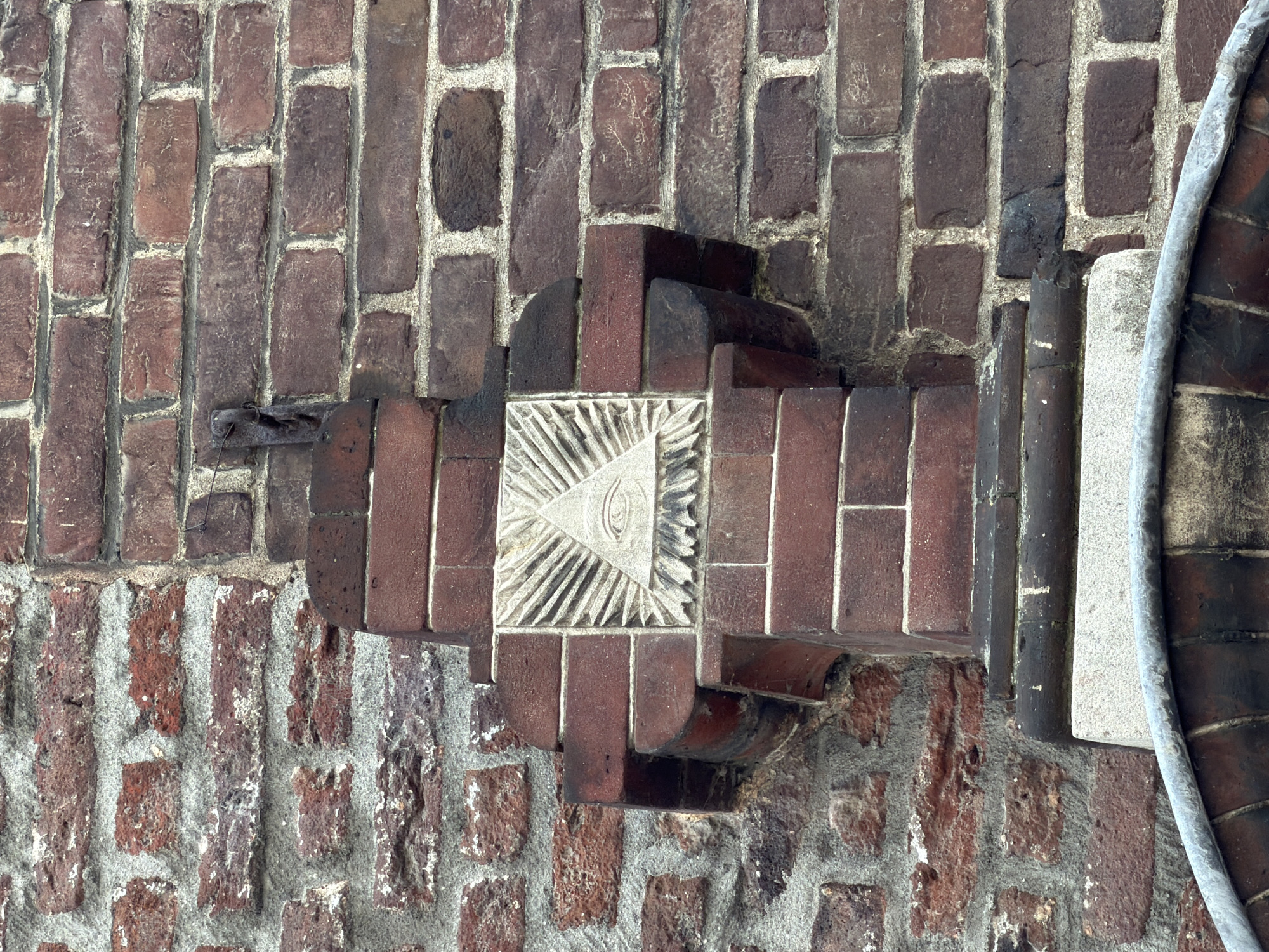 This is a close-up, eye-level, outdoor shot of a brick wall with a symbolic carving. The wall is constructed of red and darker red bricks with visible mortar. The carving, which is centrally located, is set into a lighter colored stone square and depicts an all-seeing eye within a triangle, surrounded by rays of light. This symbol is often associated with Freemasonry. The carving is mounted on a brick architectural element that juts out from the wall. The environment appears to be aged, with some weathering visible on the bricks and the carving. The lighting suggests it is daytime. The image captures a detail on the facade of the Munsterkerk in Roermond, Netherlands.