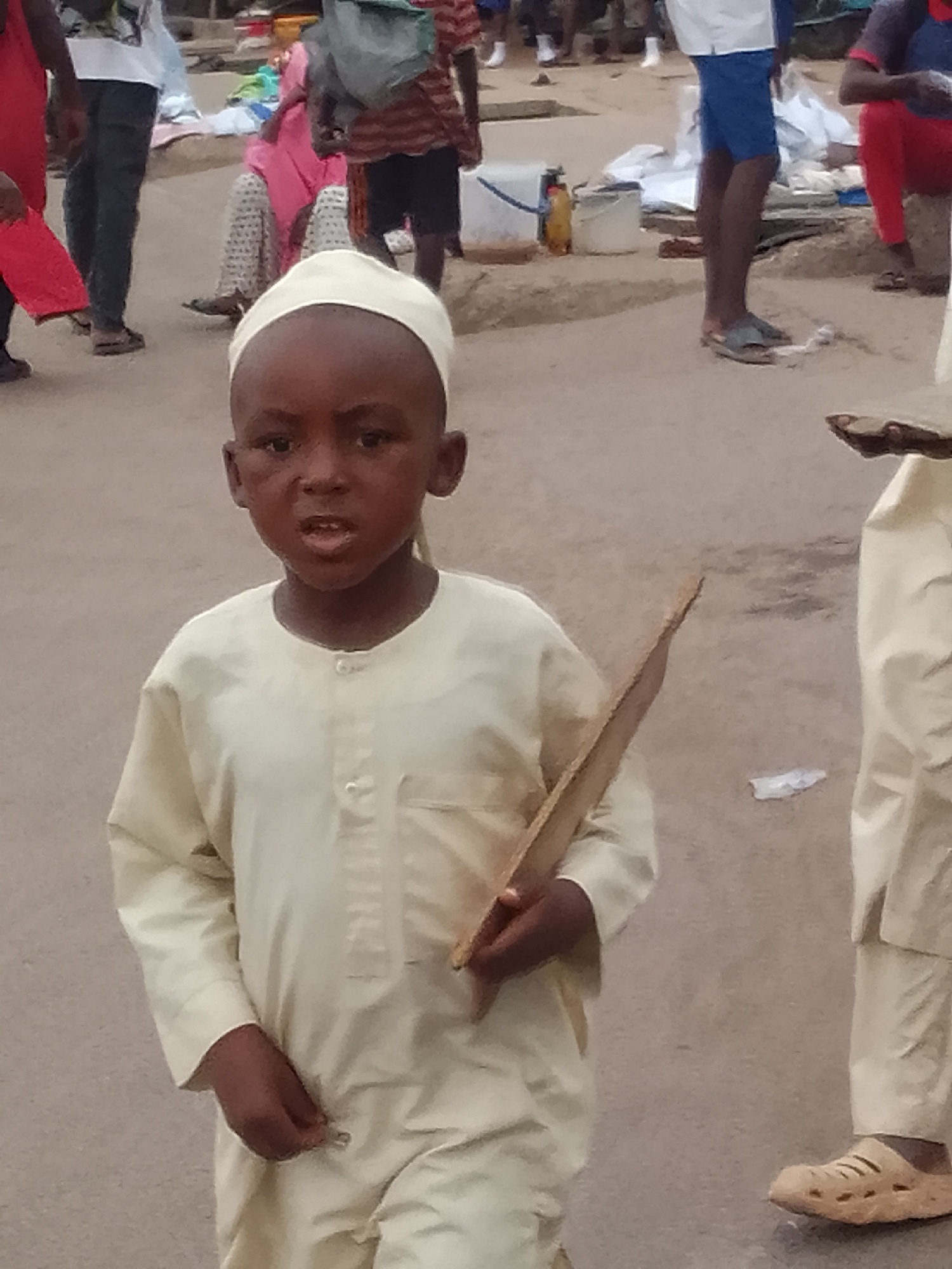 A young boy wearing a light-colored traditional outfit and a white cap stands on a dusty, outdoor market ground. He holds a flat, wooden object in his right hand. The boy's mouth is slightly open. In the background, other people are visible, some wearing colorful clothing. The environment appears to be a bustling marketplace. The context suggests this scene is in Pantame, Nigeria, a known location for vibrant market activities.