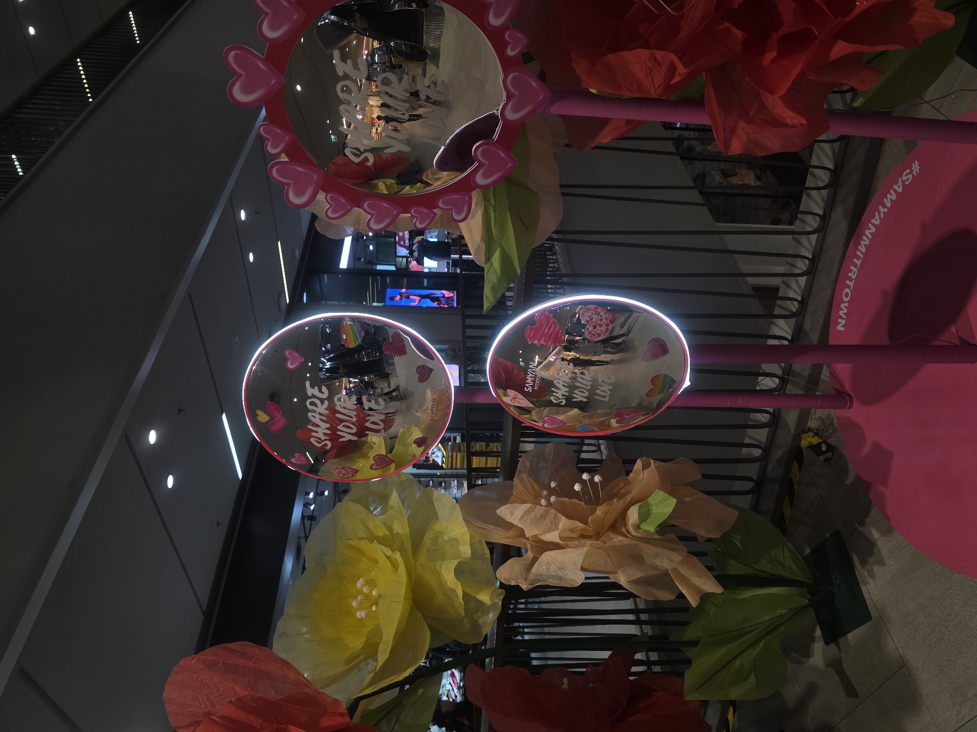 This media file captures a vibrant, love-themed decorative installation located within the Samyan Mitrtown shopping mall in Bangkok, Thailand. The scene is characterized by bright artificial lighting and a lively color palette primarily featuring pink, red, yellow, and orange.
The central subjects are several decorative mirrors, including a large heart-shaped mirror framed with pink hearts and multiple round mirrors, some encircled by glowing white rims. Text visible on these mirrors includes "SHARE YOUR LOVE" and "SAMYAN." Large, artificial paper or fabric flowers in yellow, orange, and red hues, along with green foliage, adorn the display. A prominent pink circular base on the right side of the image clearly displays the hashtag "#SAMYANMITRTOWN" in white, serving as a key location identifier for this Bangkok mall.
The reflective surfaces of the mirrors offer glimpses of the surrounding mall environment, showing escalators, overhead lighting fixtures, other store displays, and the indistinct reflections of people, suggesting an active public space. One mirror also reflects what appears to be a rainbow-colored item.
The installation suggests an event or seasonal celebration, possibly Valentine's Day or a general 'love' campaign, designed to be visually appealing and potentially interactive for visitors. The overall atmosphere conveyed is cheerful and celebratory, encouraging positivity. The indoor setting precludes determining the time of day.