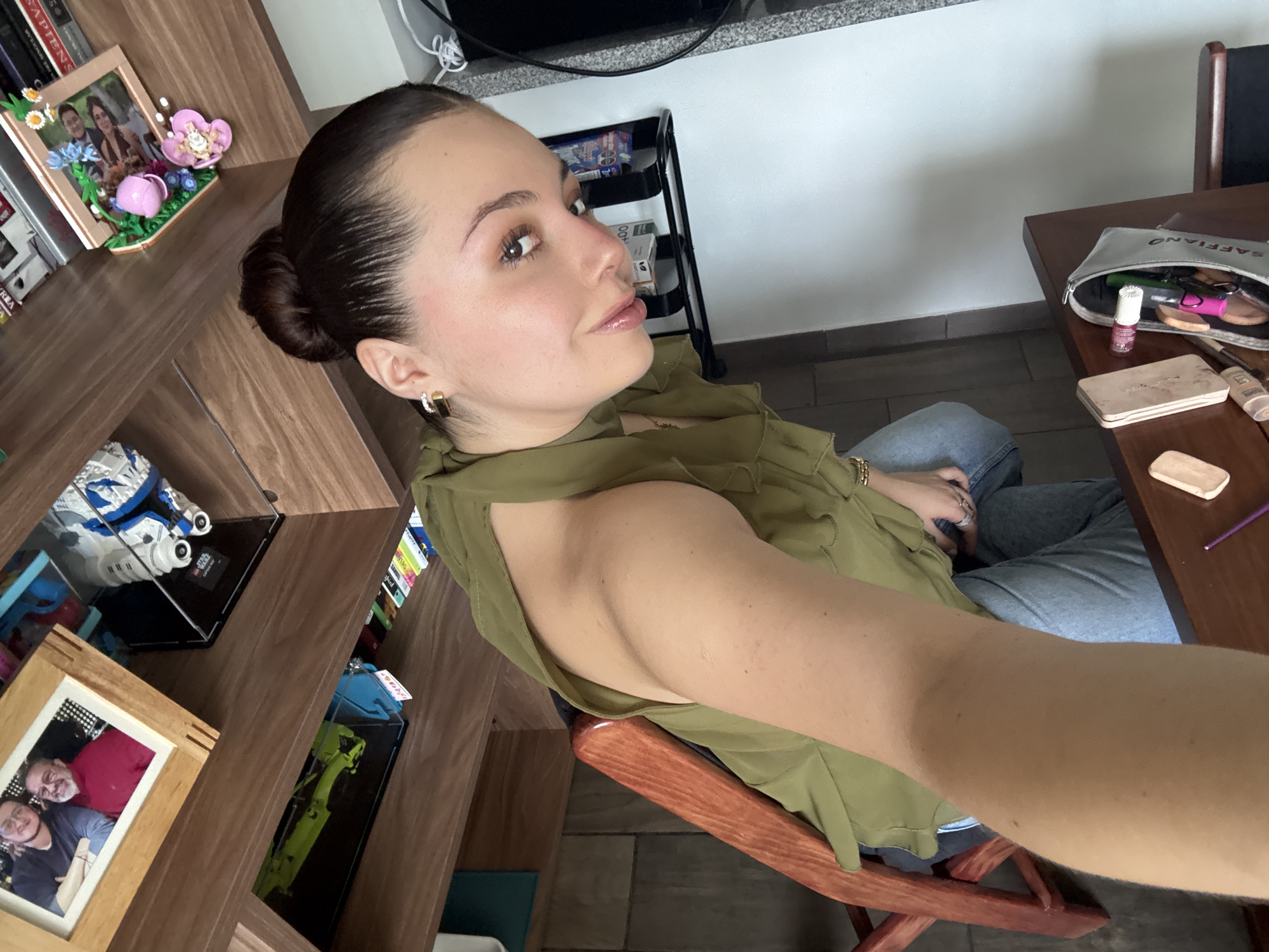 A woman with dark hair styled in a bun is seated on a wooden chair in an indoor setting. She is wearing a green top with a shoulder cutout and blue jeans. Her skin has a fair tone, and she is wearing earrings. She is looking off to the side with a slight smile.
To the left of the woman, there is a brown wooden shelf filled with various items, including framed photographs and LEGO sets in clear cases. Behind her is a black shelving unit with boxes. To her right is a brown wooden table with a makeup bag and several makeup items scattered on top. The floor appears to be tiled.
The location is potentially a residence in Mexico City, Mexico, but there is no evidence in the image to confirm this.