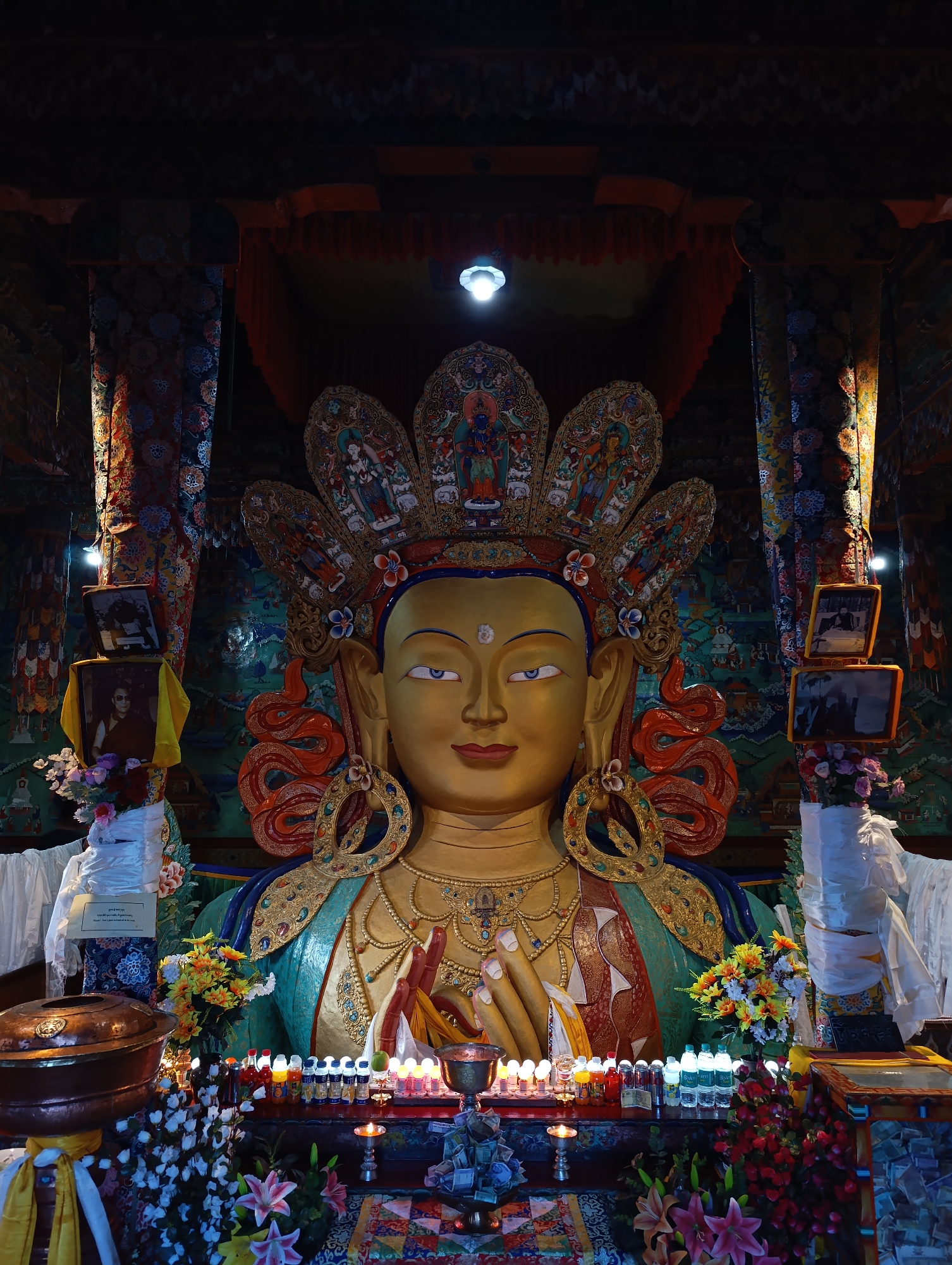 The image shows a close-up, frontal view of a large, golden Buddha statue within a dimly lit, ornate chamber. The statue is adorned with a large, elaborate headdress featuring multiple tiers of miniature paintings and decorative elements. Its face is serene, with downcast eyes and a gentle smile. The statue wears a multi-layered robe, primarily teal and red, with gold embellishments.
In front of the statue, a low altar is set up, covered with various offerings. These include numerous small bottles, likely containing liquids, arranged in rows, interspersed with lit candles and small lamps. Several bouquets of colorful artificial flowers are placed on either side of the altar and around the base of the statue. White fabric is draped around the base of the statue and on pillars flanking the altar.
To the left and right of the statue's head, framed pictures are visible, attached to the ornate interior walls. The walls themselves are covered with intricate, colorful murals depicting various figures and scenes. The ceiling of the chamber is dark and features decorative wooden beams and hanging fabrics. The overall atmosphere is one of reverence and devotion, illuminated by the soft glow of the candles and the statue's golden hue.