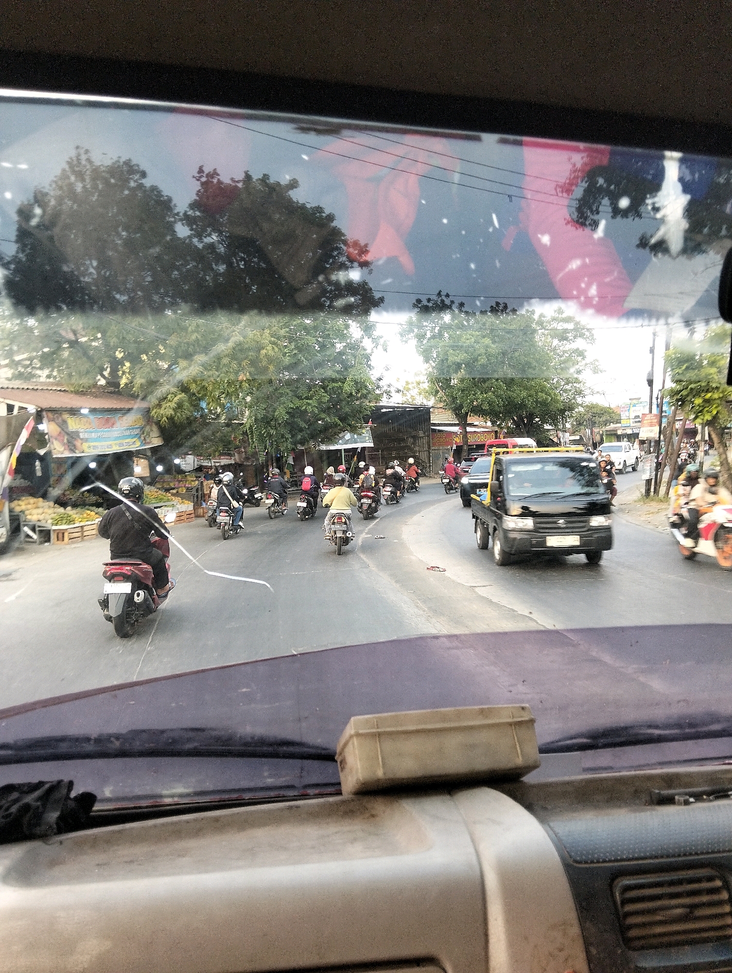 The image captures a street scene in Semarang, Indonesia, as viewed from inside a vehicle, likely a truck given the dashboard detail. The perspective is from behind the windshield, which shows reflections of trees and some reddish-pink material, possibly banners or cloth. The road is paved and appears to be a typical urban street with multiple lanes.
The scene is busy with traffic, predominantly motorcycles. Several individuals are riding motorcycles in various directions. One person in the foreground on a red scooter is wearing a black jacket and helmet. To the right and further down the road, a black truck with a flatbed is prominent, suggesting it's a commercial vehicle. Other vehicles, including what appears to be a white SUV, are also present.
Along the sides of the road, there are buildings with awnings, some of which appear to be market stalls selling produce like fruits. Trees line the street, providing some shade. Power lines are visible overhead, a common feature in urban Indonesian landscapes. The overall impression is of a lively, active street environment in a tropical climate, typical of Semarang. The condition of the dashboard and windshield suggests the vehicle is utilitarian rather than luxurious.
