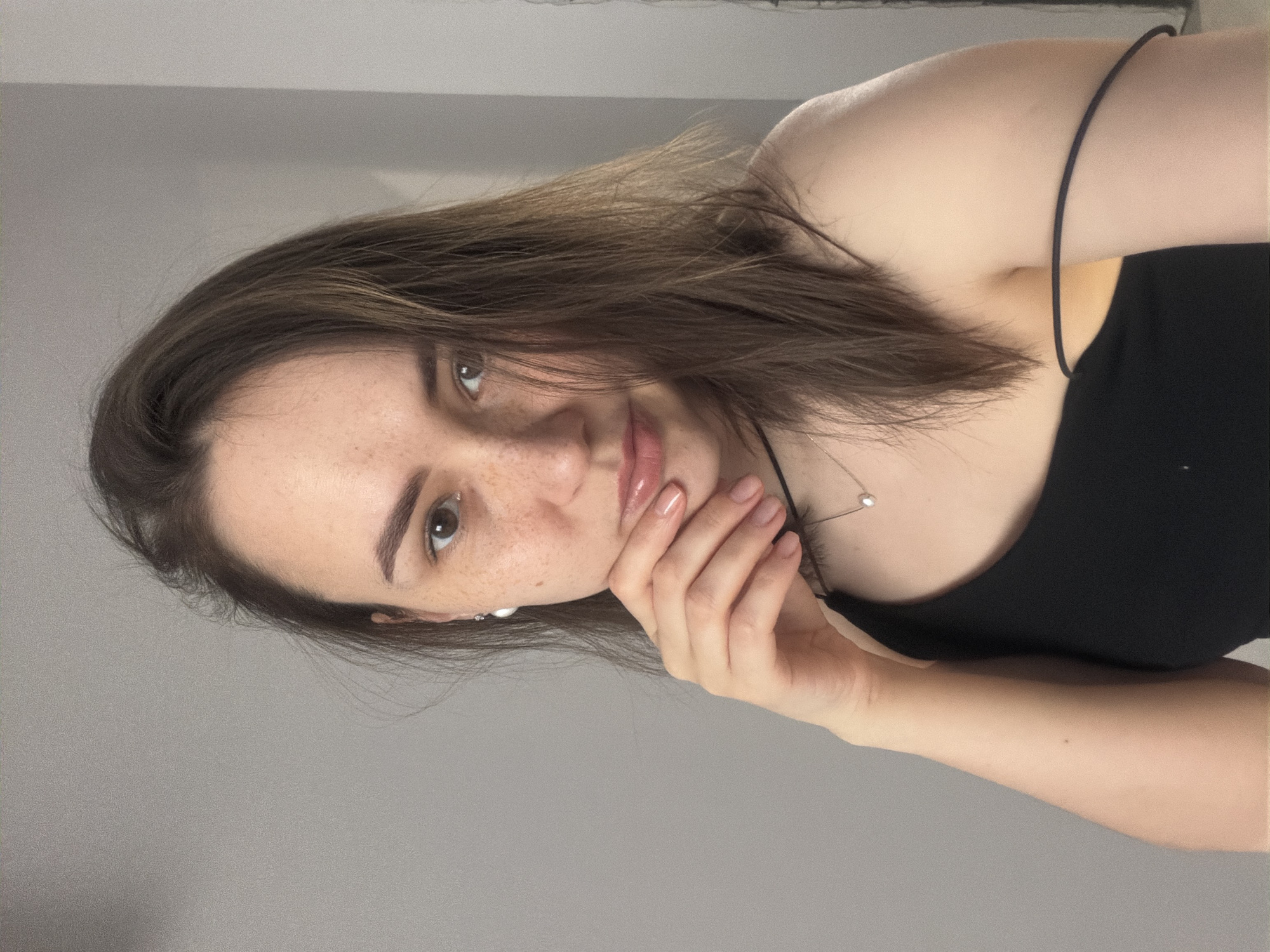 A young woman poses for a close-up selfie, her left cheek resting on her hand. She has fair skin, brown hair, and visible freckles on her face. She is wearing a black top with thin straps and a delicate necklace. A pearl-like earring is visible in her left ear. The background is a plain grey wall. There is no indication of this image being taken in the city of Chaikovsky, Russia.