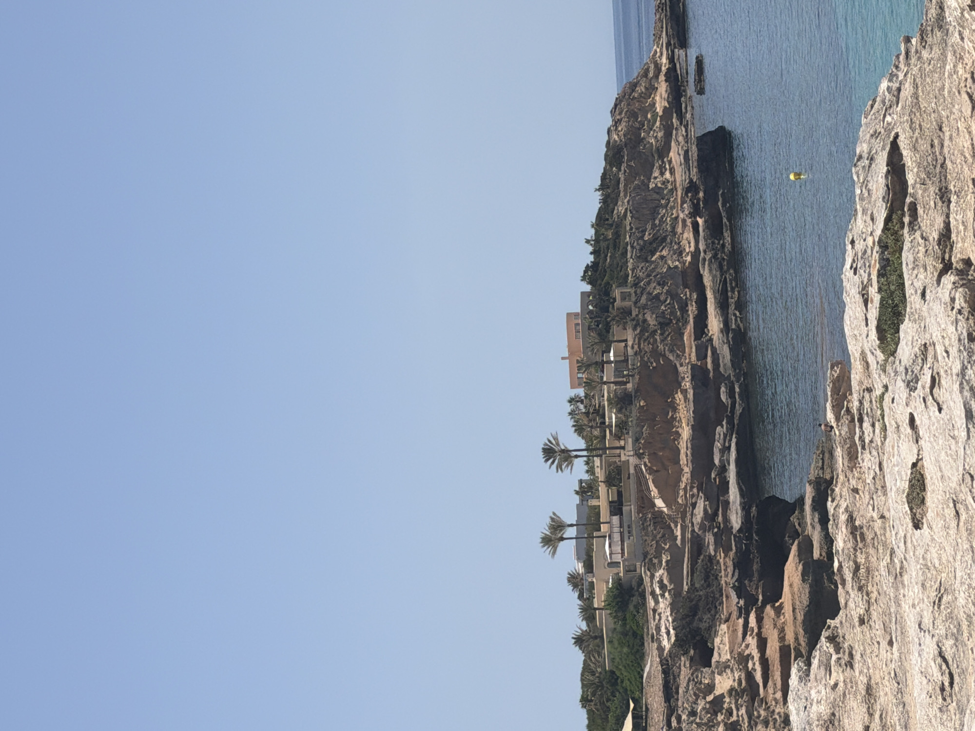 The image captures a coastal scene in Sant Josep de sa Talaia, Spain, under a clear blue sky. The foreground is dominated by rugged, light-colored rock formations that slope down towards the sea. In the middle ground, a rocky cliff juts out into the calm, blue water. On top of this cliff, there are several buildings, some with flat roofs and light-colored facades, interspersed with palm trees and dense green vegetation. A larger building with an orange roof is visible on the far right. The water appears tranquil, with a single yellow buoy floating in the sea. In the lower right portion of the image, a lone person stands on the rocky shore, facing away from the camera. The overall impression is of a sunny, Mediterranean coastal landscape.