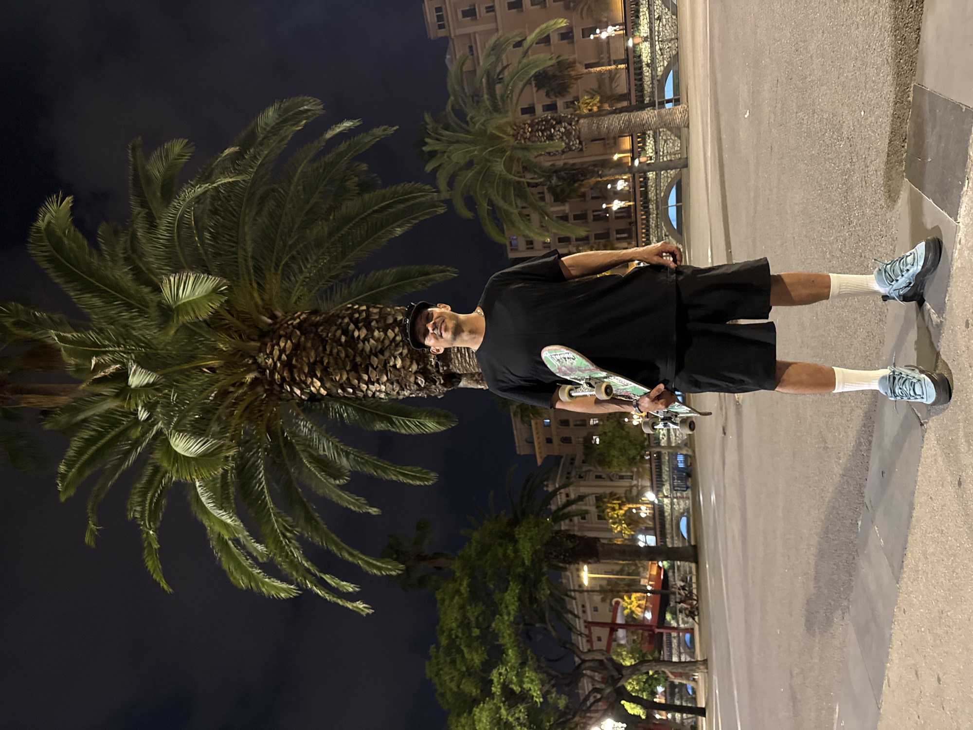 A man stands in an urban environment at night, holding a skateboard. He is wearing a black t-shirt, black shorts, white socks, and blue sneakers, and he has a baseball cap on his head. Palm trees are prominent in the background and to the left, with illuminated buildings visible behind them. The setting is a street in Barcelona, Spain, evident by the characteristic palm trees lining the streets and the style of the architecture. The man is standing on the sidewalk, next to a street illuminated by streetlights. The street surface is asphalt.