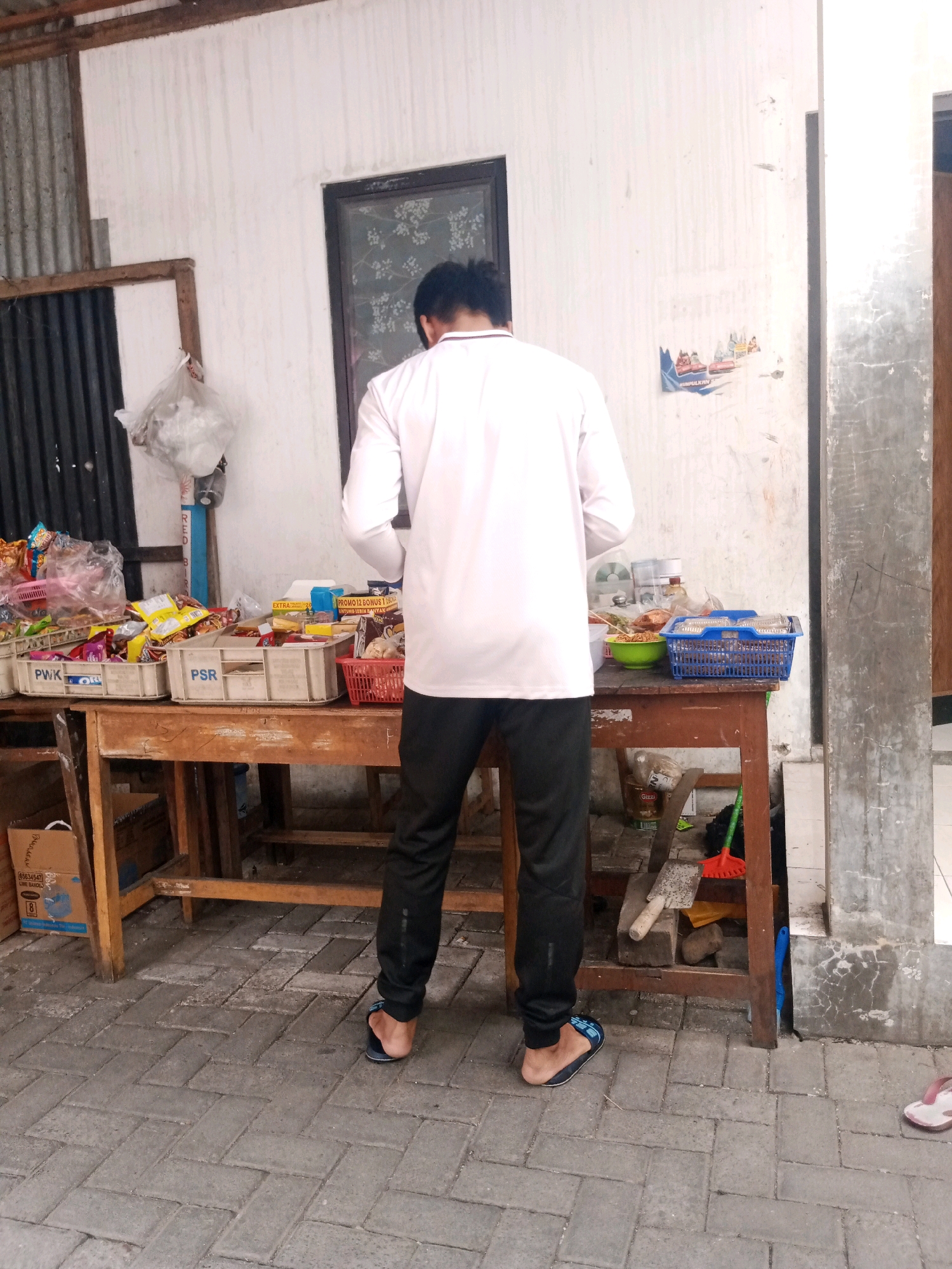 A man wearing a long-sleeved white shirt and black pants stands facing away from the camera, looking at a table laden with snacks and groceries. He is wearing blue and black flip-flops. The table is made of wood and is supported by wooden legs. Behind the table, a white wall with some water stains and a window with a floral pattern is visible. To the left of the table, there's a corrugated metal fence. The ground is paved with interlocking bricks. To the right of the table, a broom and a shovel are leaning against the wall. The scene appears to be an outdoor stall or shop, likely in a residential area of Surakarta, Indonesia, given the architectural style and the presence of common Indonesian packaged goods.
