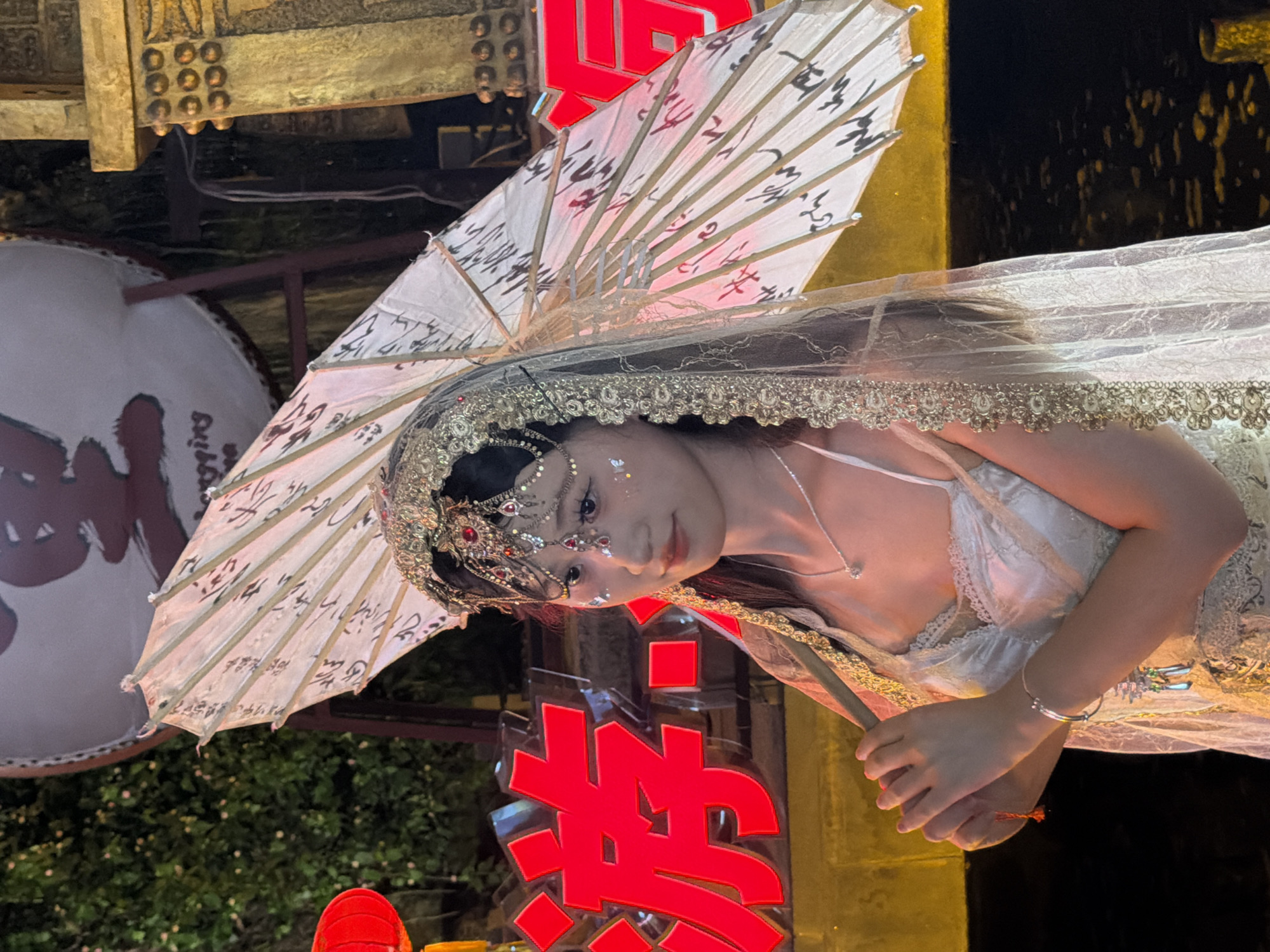 A young East Asian female stands outdoors at night, holding a closed white umbrella with black calligraphy. She wears an elaborate headdress adorned with red jewels and gold accents, and a white veil with gold trim. Her attire includes a white lace-trimmed camisole and a matching dress. A delicate necklace and a bracelet with charms are visible. Glitter is applied to her face, around her eyes and cheeks. Behind her, large red Chinese characters are illuminated. To her right, a golden structure with decorative circular elements is visible. In the background, a large drum with Chinese calligraphy is present, and some greenery is to the left. The scene is likely a night market or festival, possibly in the Yuzhong District of China, given the cultural elements and signage.