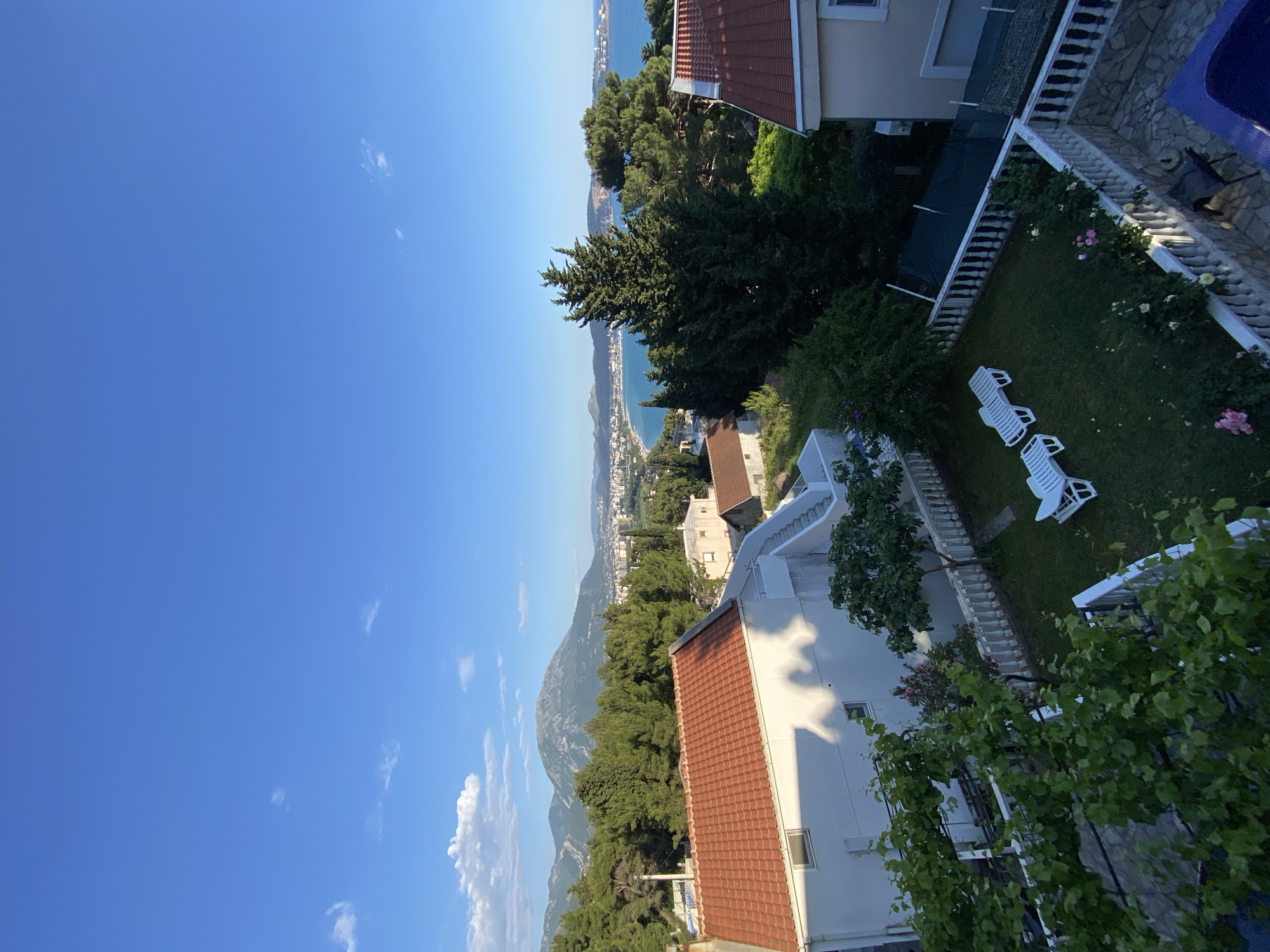A landscape view from a high angle features the city of Šušanj, Montenegro. The scene includes a blue sky with a few scattered clouds, a mountainous region, and the Adriatic Sea in the background. Residential buildings with red-tiled roofs are visible amidst lush green trees and vegetation. A private garden with white lounge chairs and a white balustrade is present in the foreground.