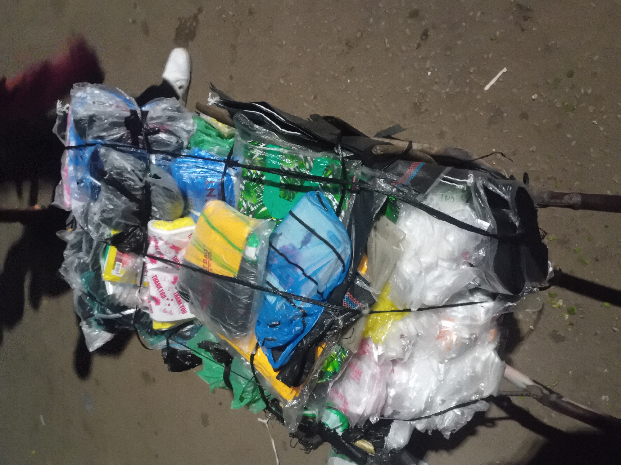 This image from Bukuru, Nigeria, captures a large, tightly bundled load of various items, predominantly consisting of colorful plastic bags and packaged goods. The bundle is secured with dark straps or cords, holding together a diverse collection of items in transparent, blue, green, yellow, white, and black plastic packaging.
Two dark wooden poles extend horizontally from either side of the bundle, suggesting it is being prepared for transport or actively carried. A person's leg and foot, clad in a white shoe, are partially visible on the upper left, and a human shadow is cast on the ground to the lower left, indicating a person is near or involved in moving the load. The ground is dark and uneven, possibly dirt or a worn path, illuminated by artificial light, suggesting the photo was taken at night or during very dim light conditions.
Visible text includes the phrase "THANK YOU" repeated in red on several white plastic bags, and a partial word "BAG" on a yellow bag. The scene depicts a common method of transporting goods in many regions, emphasizing resourcefulness in conveyance.