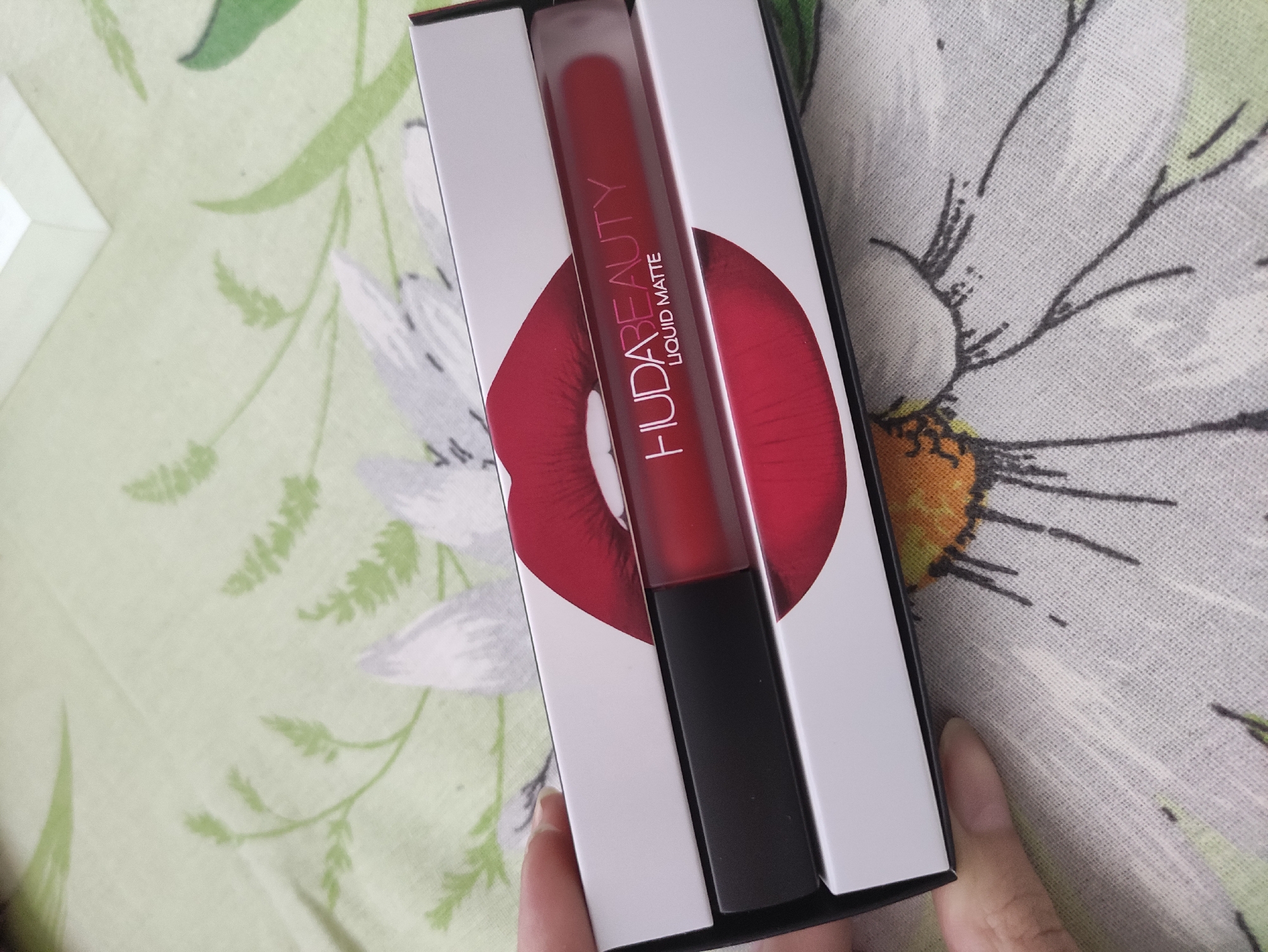 A person's hand is holding a rectangular box of Huda Beauty liquid matte lipstick. The box is white with a graphic of red lips and the Huda Beauty logo printed on it. The lipstick tube itself is visible inside the box, with a black cap and a clear tube showing the red lipstick. The box is placed on a light green fabric with a floral pattern.