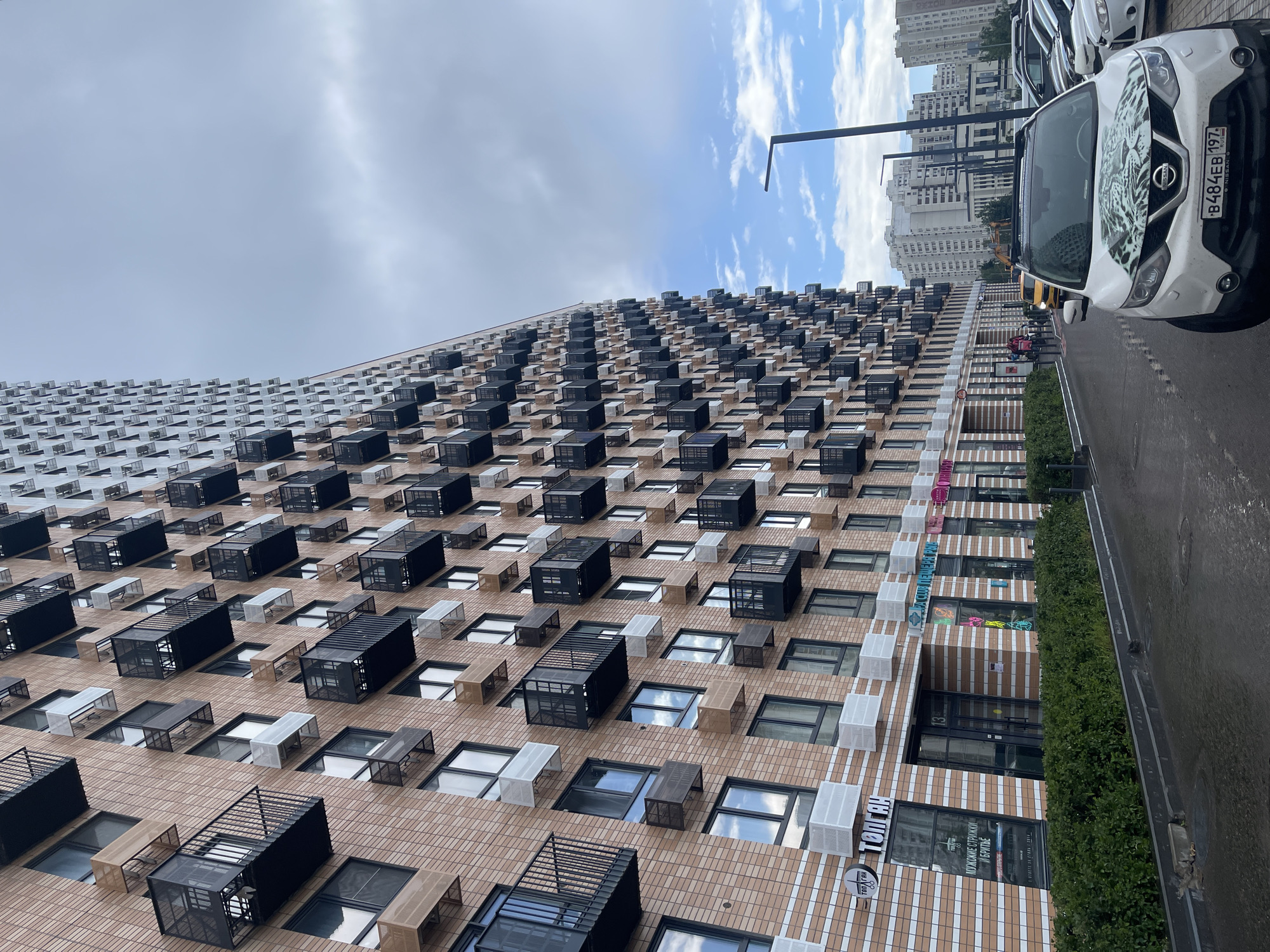 The media file captures a section of a modern residential building in Moscow, Russia, under a cloudy sky. The building features a facade with a grid-like pattern of numerous small, dark-colored, square balconies or window enclosures, interspersed with lighter colored, rectangular window frames. The repetition of these architectural elements creates a striking visual rhythm across the building's exterior.
In the foreground, a white Nissan SUV is parked along the side of the building, its hood decorated with a black and white graphic of a big cat. The car's license plate, B484EB 197 RUS, is clearly visible. To the right of the car, a lush green hedge runs along the pavement, bordering the building. Further down the street, other buildings and a yellow excavator are visible in the background, suggesting ongoing construction or urban development.
Several signs are visible on the building's lower levels. One sign, partially obscured, appears to be for a barbershop, with text in Russian including "КУХОННЫЕ СТРИЖКИ И БРИТЬЕ" which translates to "Kitchen haircuts and shaving," likely a playful or stylized business name. Another sign reads "ТОП ГАН." The sky is partly cloudy, indicating it is likely daytime. The overall impression is one of contemporary urban architecture and city life.