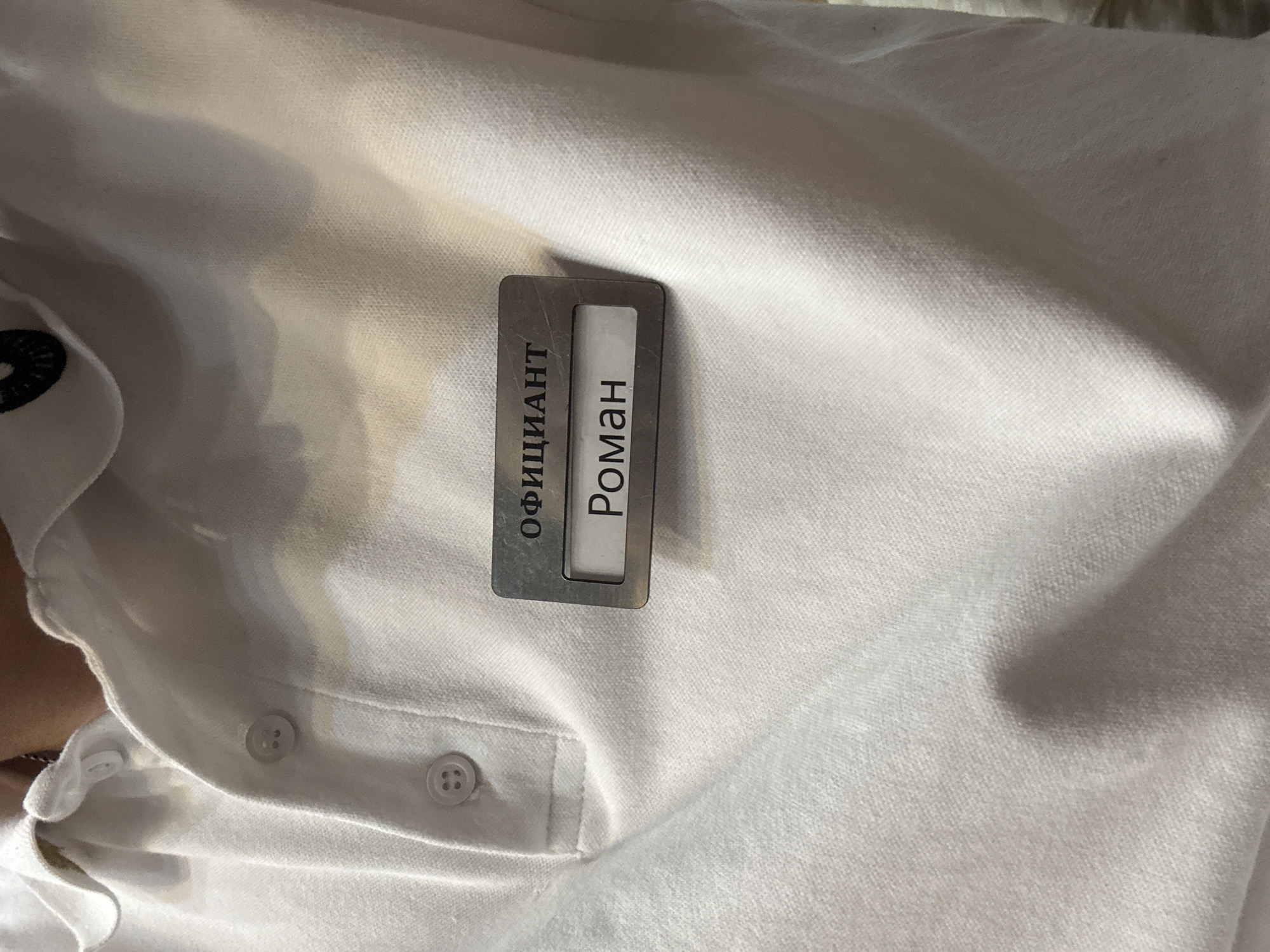 A close-up image captures a metallic name tag affixed to a white polo shirt. The shirt features a piqué knit fabric and visible white buttons on its placket. The name tag is rectangular with a brushed metal frame, holding a white insert with black Cyrillic text. The top word reads "ОФИЦИАНТ" (OFITSIANT), translating to "WAITER." Below it, the name "Роман" (ROMAN) is displayed. No person's face or distinct body parts are visible, only the upper torso area of the shirt. The lighting suggests an indoor setting. The scene is situated in Birobidzhan, Russia.