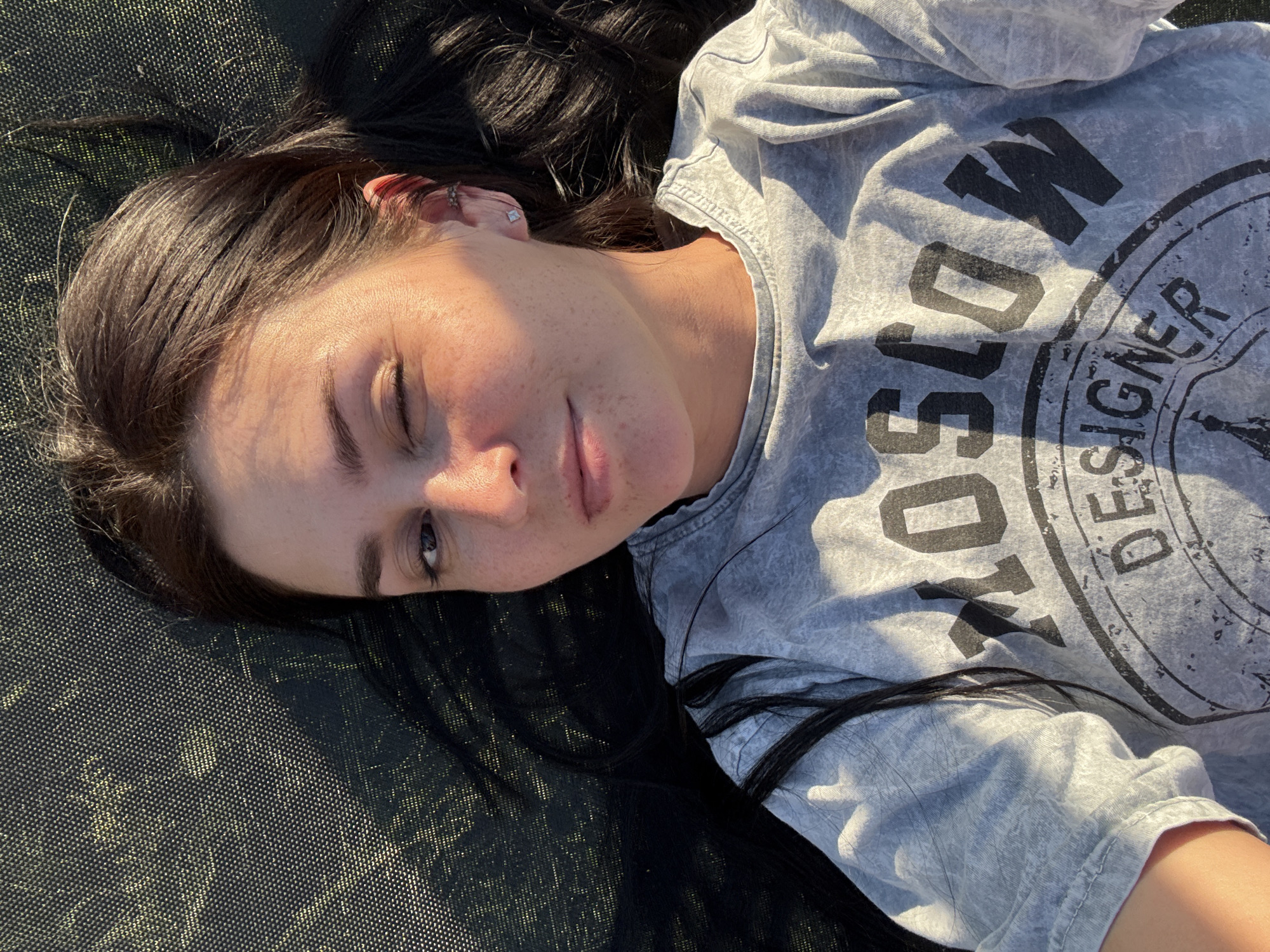 A young woman with dark hair is lying down outdoors, looking up towards the camera. She is wearing a grey t-shirt with "MOSCOW DESIGNER" printed on it in large, bold black letters. The woman's face is partially illuminated by the sun, casting shadows and highlighting her features. She has freckles across her nose and cheeks and is winking. Her hair is spread out around her head. The surface she is lying on appears to be a dark, textured material, possibly a trampoline or outdoor mat, with a slightly metallic sheen in places. The lighting suggests it is daytime, with the sun casting long shadows. The overall mood is relaxed and casual.