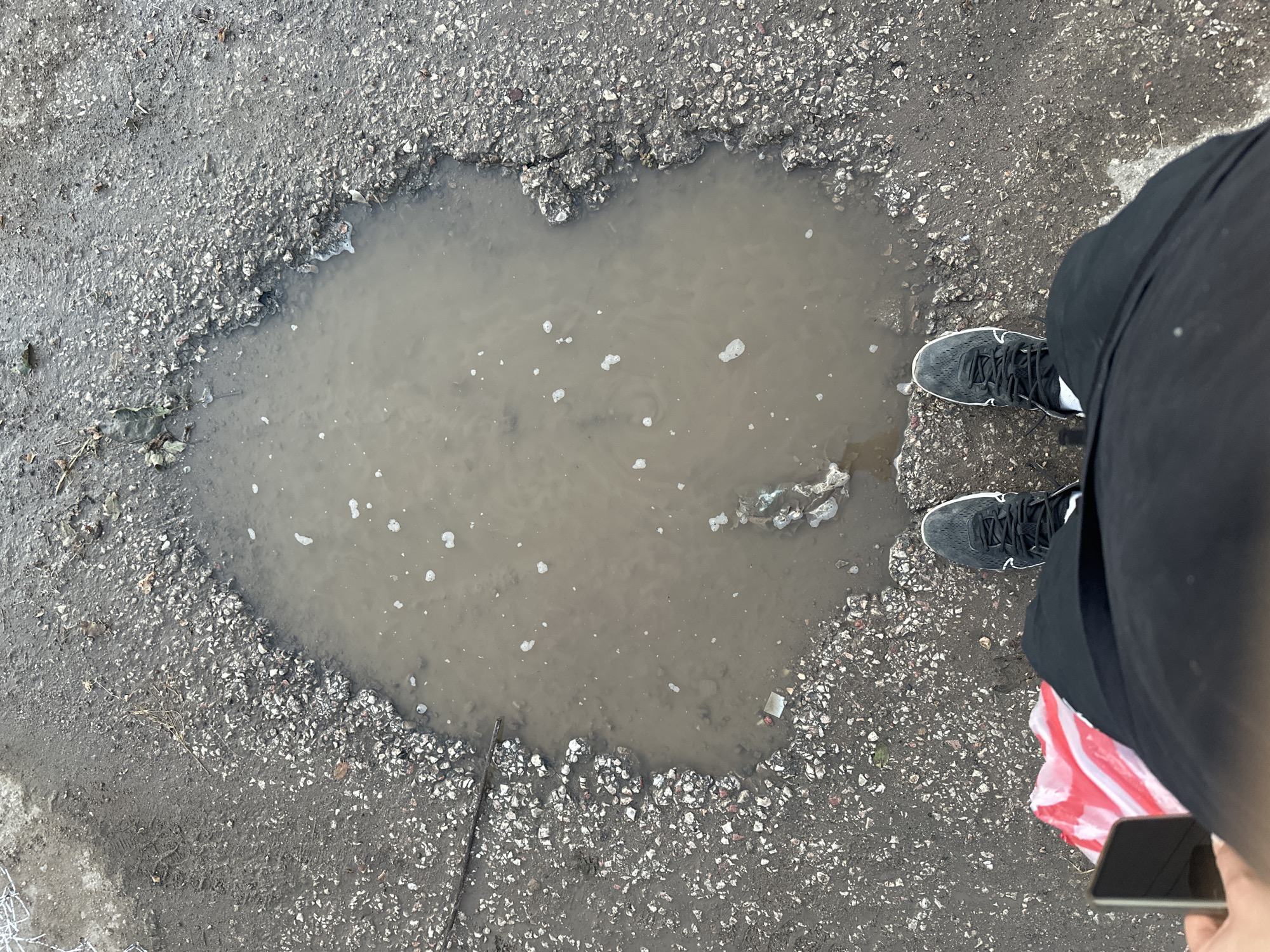 The media file shows a person standing next to a large pothole filled with murky water on an asphalt surface. The person is wearing black pants and black sneakers with white accents. A red and white striped bag is partially visible, held by the person. The person is holding a smartphone in their right hand, angled downwards. The pothole is irregular in shape, with jagged edges of broken asphalt and gravel. The water in the pothole is brown and has several small white bubbles on its surface. The surrounding ground is also asphalt, with loose gravel and some dry leaves. A thin stick or twig lies across the edge of the pothole. The lighting suggests it is daytime, and the overcast sky implied by the lighting and the presence of the puddle hints at recent or ongoing rain. The scene appears to be an outdoor urban or suburban environment, possibly a street or pathway with poor road maintenance. There is no visible text in the image.