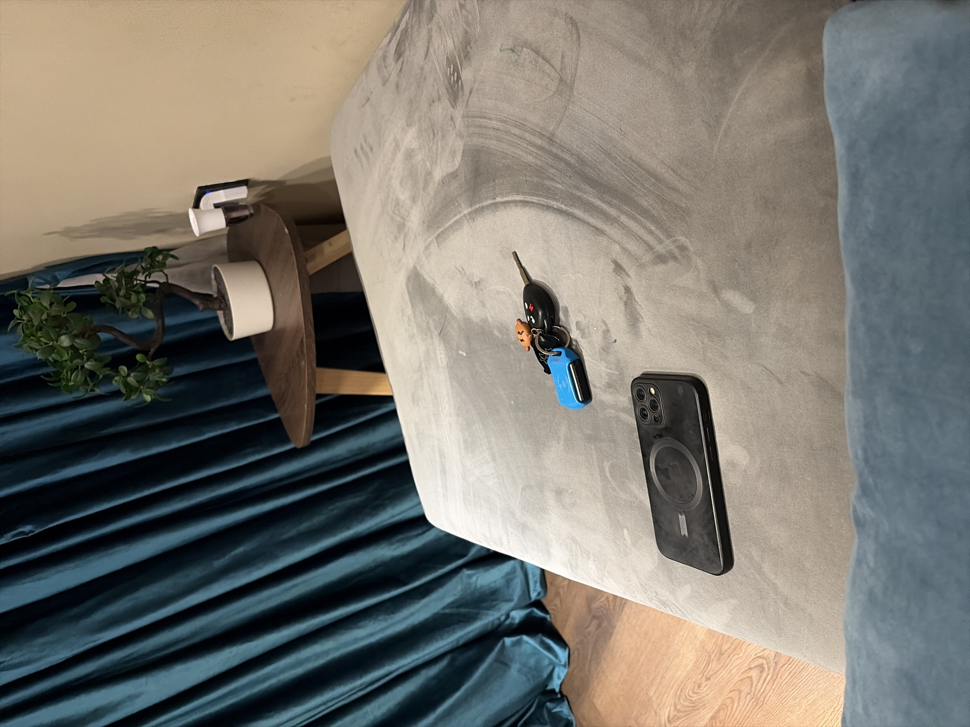 The image shows a room interior. A gray upholstered sofa dominates the foreground. On the sofa are car keys with a blue keychain and a black smartphone with a circular MagSafe charging interface. To the left of the sofa, there's a small round wooden table supporting a potted plant with green foliage. On the wall above the table is a white and black wall-mounted device. Below the table and plant is a textured teal-colored fabric. The floor adjacent to the sofa is light-colored wood. No people are visible in the scene. The location in the city of Abuja, Nigeria is undetermined.