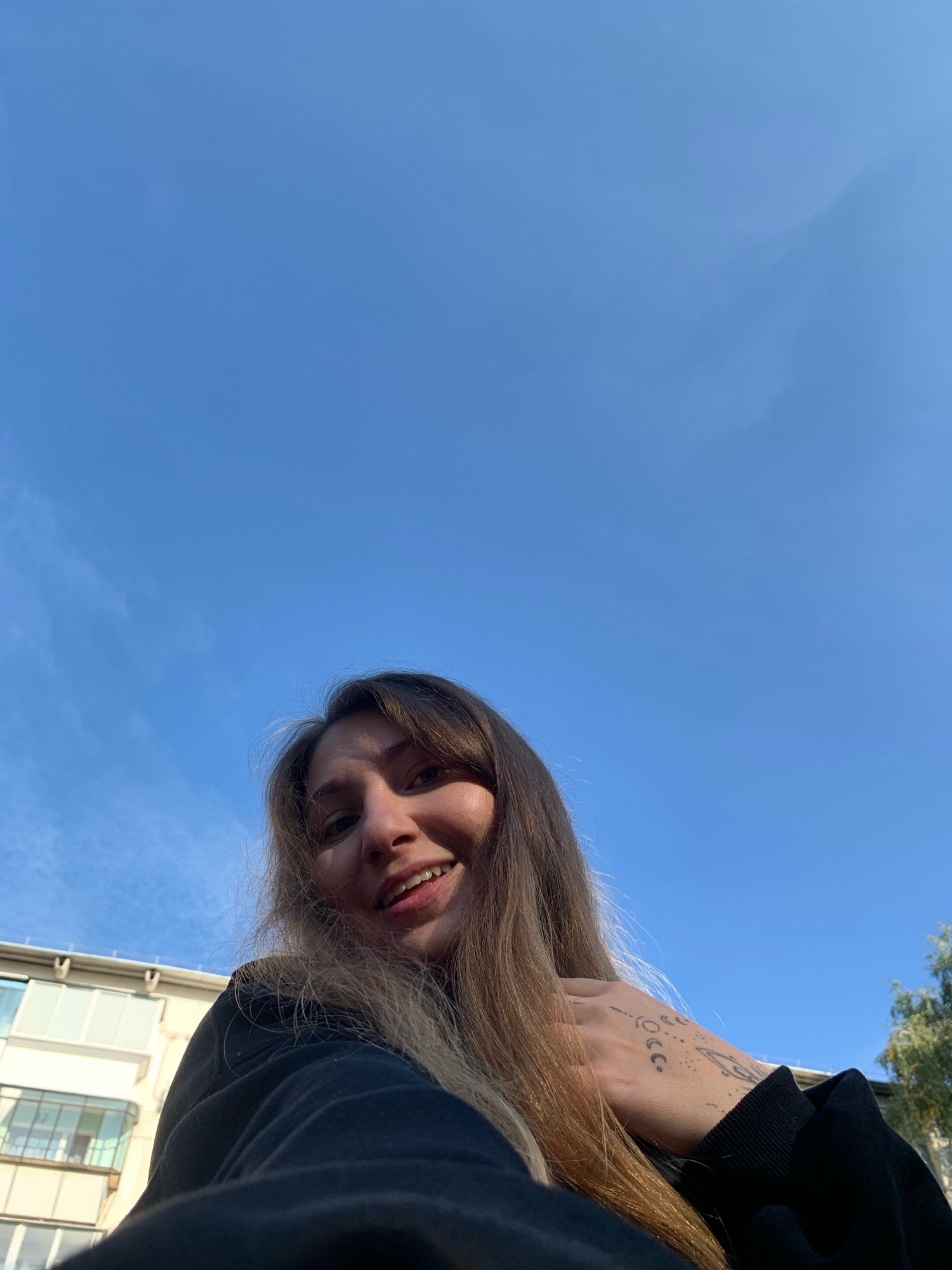 The image is a low-angle, close-up selfie taken outdoors, likely in the city of Chaikovsky, Russia, given the context. The background is a clear, bright blue sky with a few wispy clouds. In the foreground, a young woman is looking up at the camera with a smile, her face partially illuminated by sunlight. She has long, light brown hair that falls around her face. She is wearing a dark-colored hooded sweatshirt. A tattoo is visible on her left hand and wrist, featuring several celestial symbols, including moons and circles. To the left and slightly behind the woman, a portion of a building with balconies and windows is visible, suggesting an urban or suburban setting. The overall scene appears to be a casual, outdoor moment captured on a sunny day.