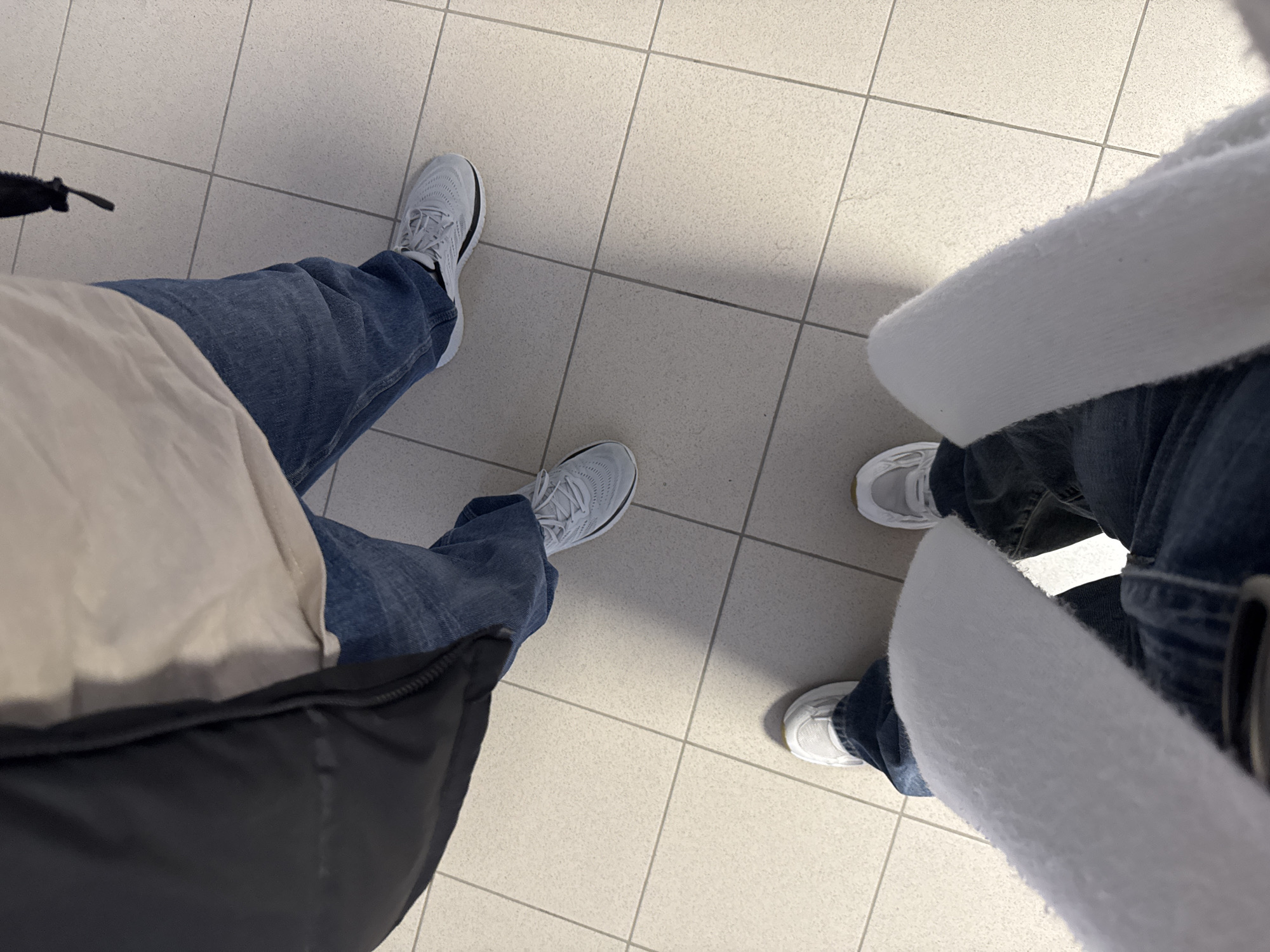 From a high-angle perspective, the image captures the lower halves of two individuals standing on a tiled floor. The tiles appear to be light-colored. The person on the left is wearing blue jeans and grey athletic shoes. A partially visible jacket with a black outer layer and a beige inner layer is present. The person on the right is wearing a white jacket and jeans and white shoes. The background is a tiled floor. No explicit location or city can be determined from the image.