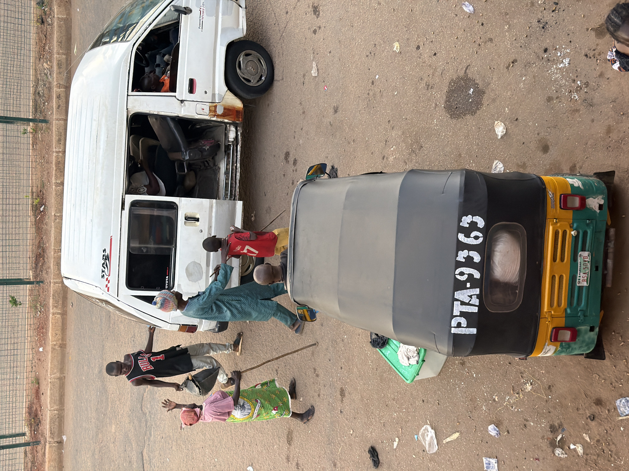 In Bukuru, Nigeria, a street scene unfolds with a white minivan and a three-wheeled auto-rickshaw as the central subjects. The minivan, bearing the "STARS" logo, has its side door open, revealing passengers inside, one in an orange shirt looking out a window. A young boy in a red jersey is near the open door, while another child in a red shirt stands by the minivan. A man in a blue kaftan and a patterned cap walks away from the minivan, holding a stick. A woman in a pink shirt and a green patterned skirt, also holding a stick, faces the minivan, her left hand raised. A young man in a black "BULLS" basketball jersey walks towards the minivan, carrying a dark bag.
The auto-rickshaw, in green, yellow, and black, is parked in the foreground, its rear visible with the license plate "RSA-50YD" and the white lettering "PTA-9363" on the back. A green plastic container with a white cloth inside is on the ground near the auto-rickshaw. The environment is a dusty, paved street with scattered debris, including plastic bottles and paper. A metal fence runs along the background, with a dirt embankment and vegetation beyond. The lighting suggests it is daytime, with shadows cast on the ground.